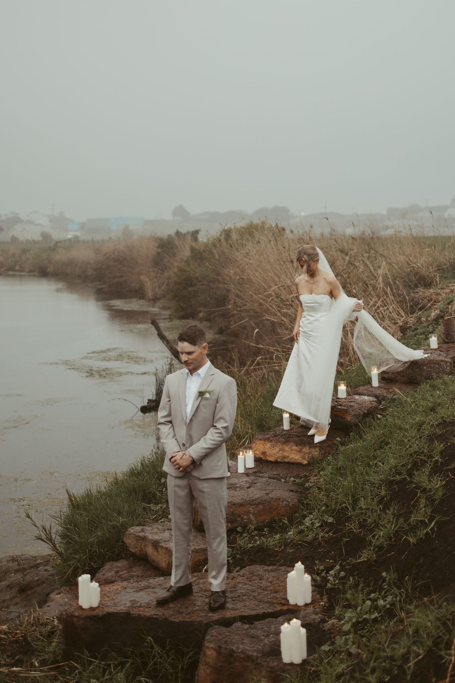 Bride in white gown and veil approaching groom on rocky outdoor ceremony site with candles.