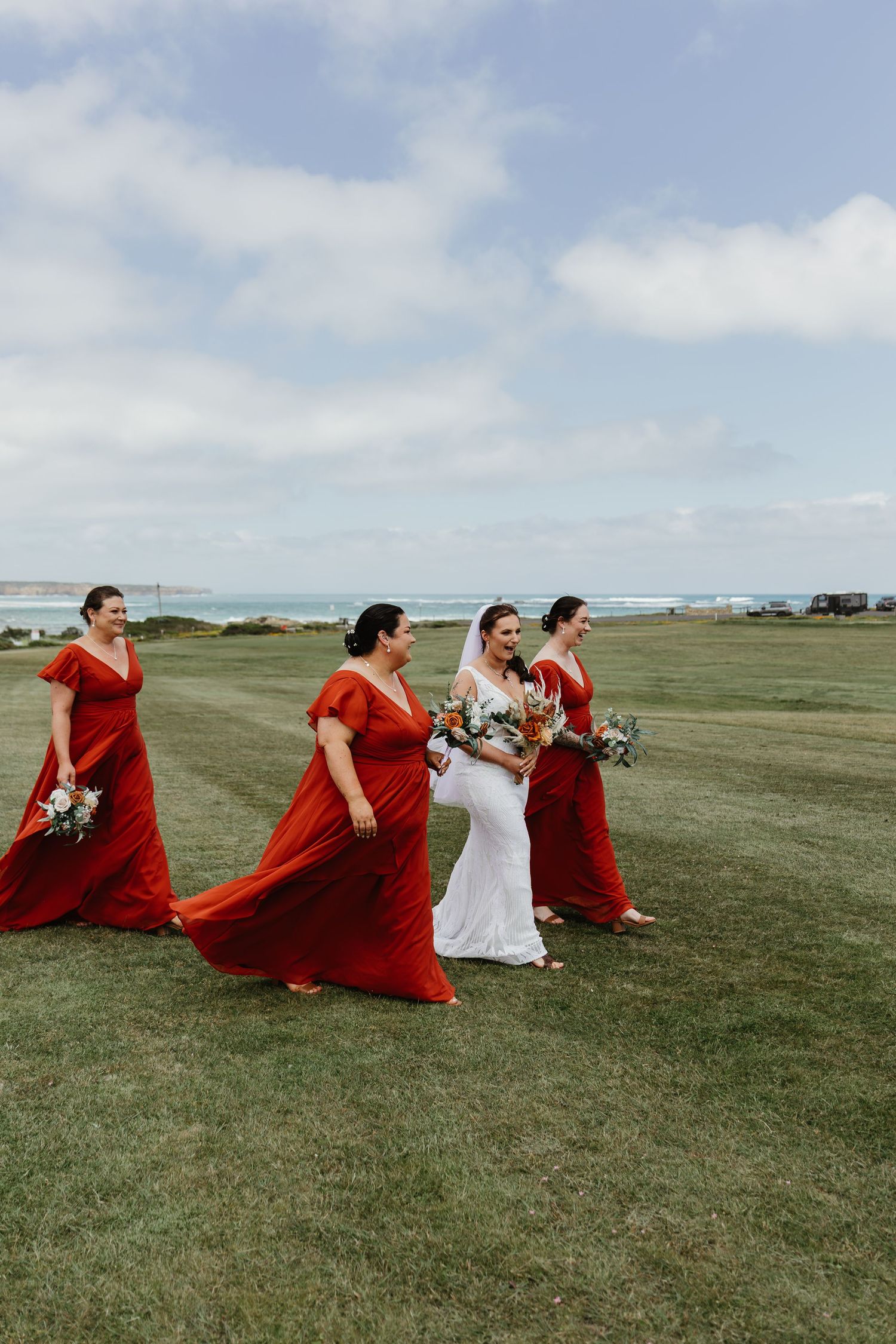 Bride in white gown walks with three bridesmaids in flowing red dresses along a coastal grassy lawn.