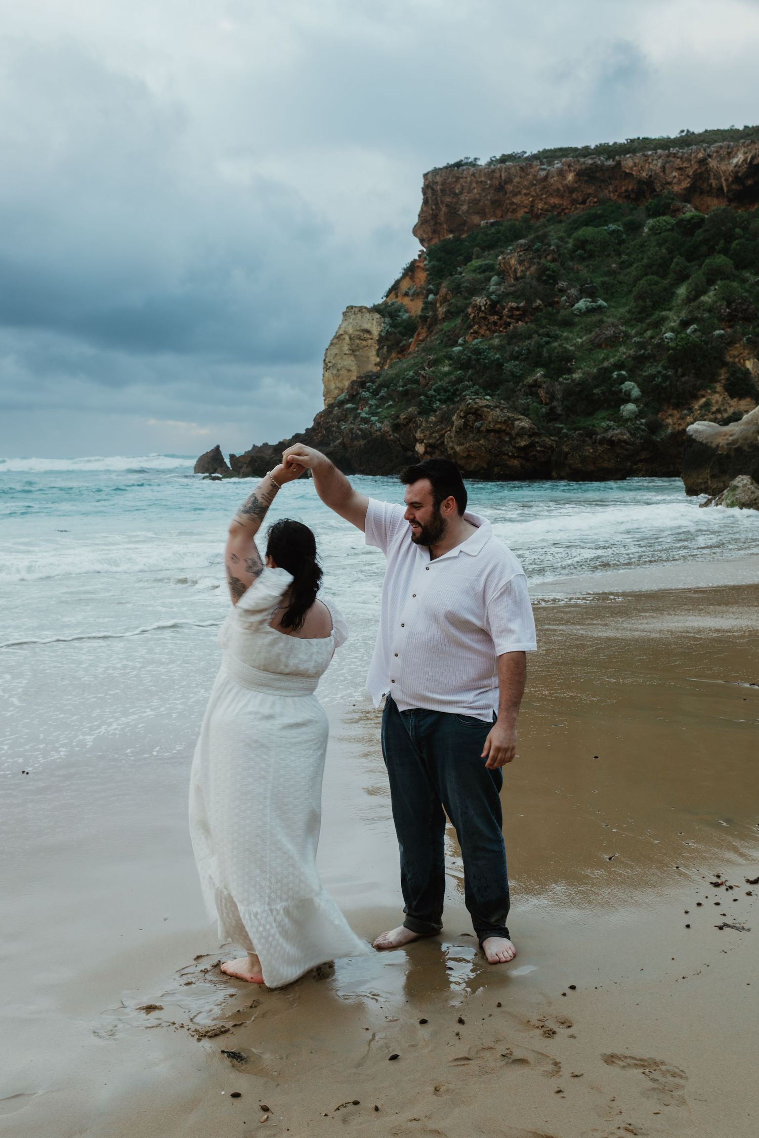 Couple dancing on a sandy beach near rocky cliffs, woman in white dress, man in white shirt and jeans.