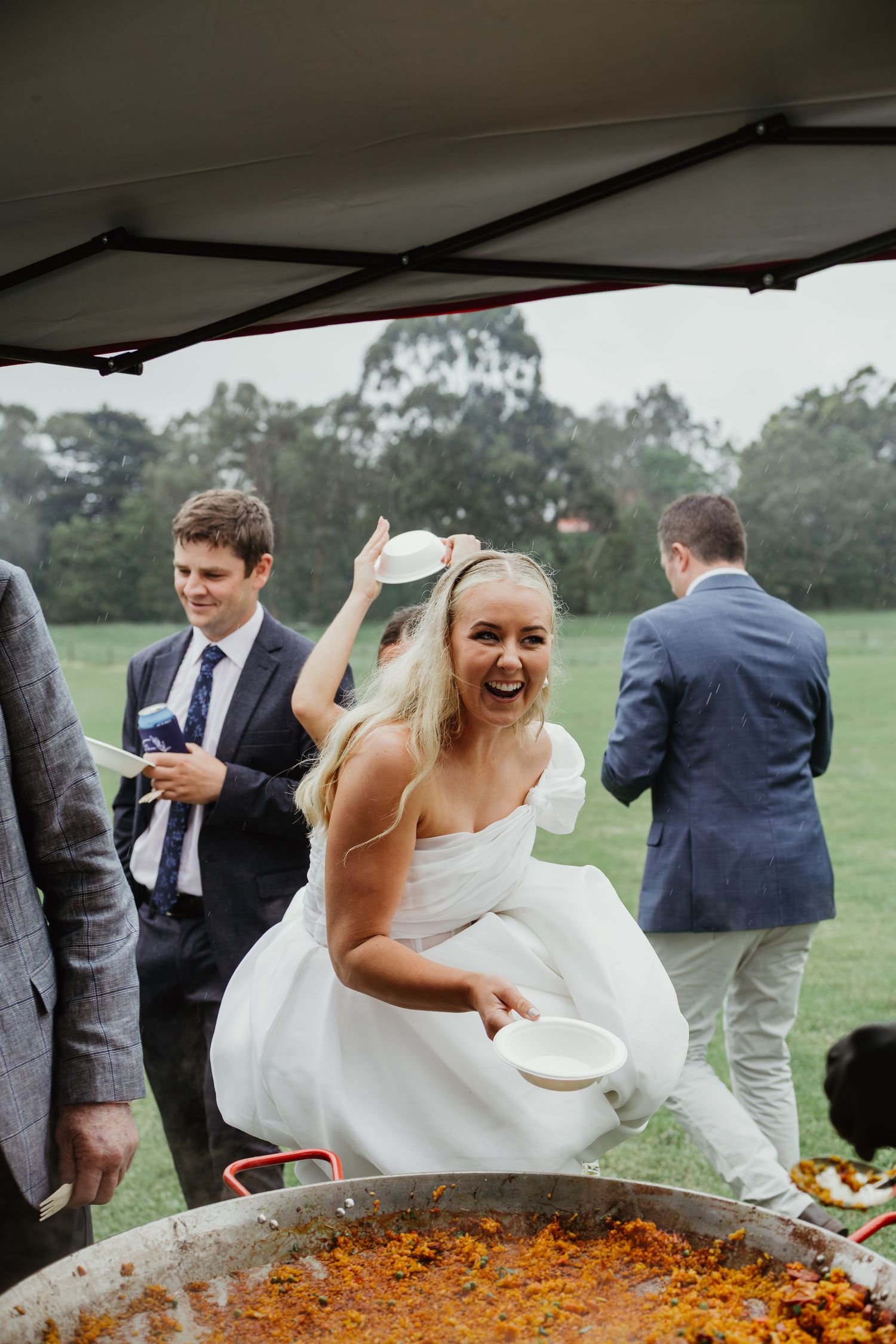 Laughing bride in white dress serving paella at outdoor wedding reception in rain.