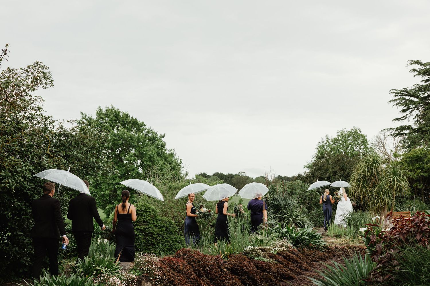 Wedding party with white umbrellas in lush garden on overcast rainy day.