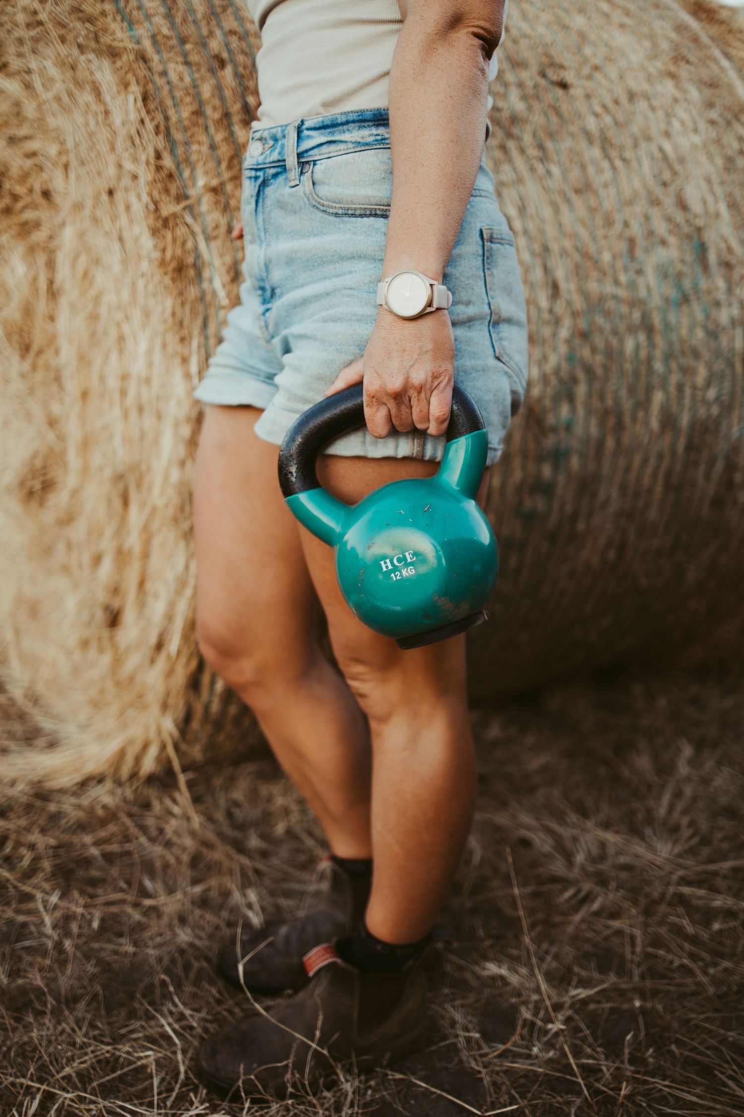 Woman in denim shorts holding a teal kettlebell outdoors near hay bales, wearing a white watch and brown boots.