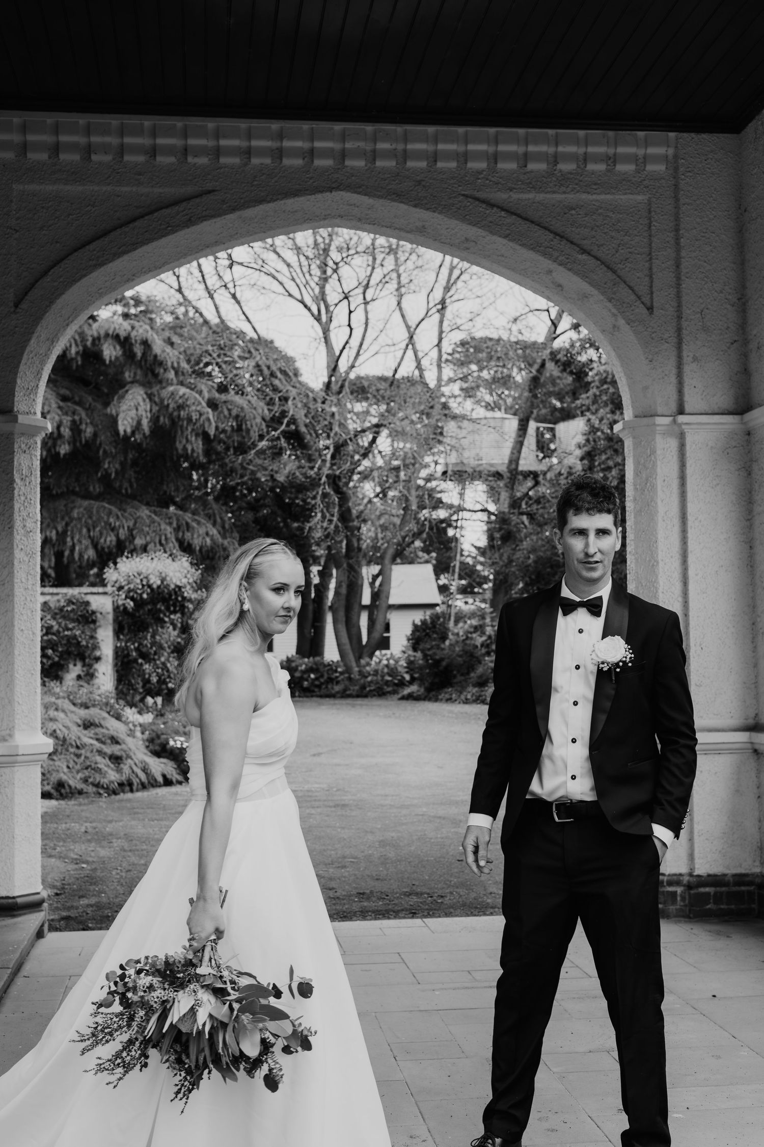 Black and white photo of bride and groom standing under an ornate stone arch in a garden setting.