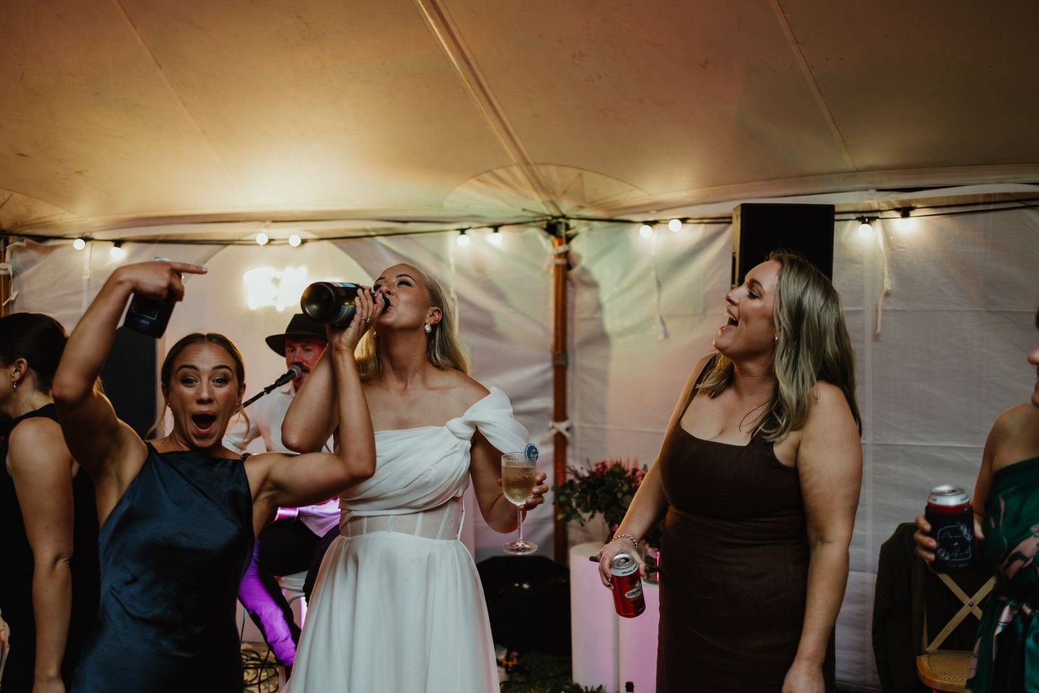 Bride and guests singing and dancing joyfully inside a warmly lit wedding reception tent.