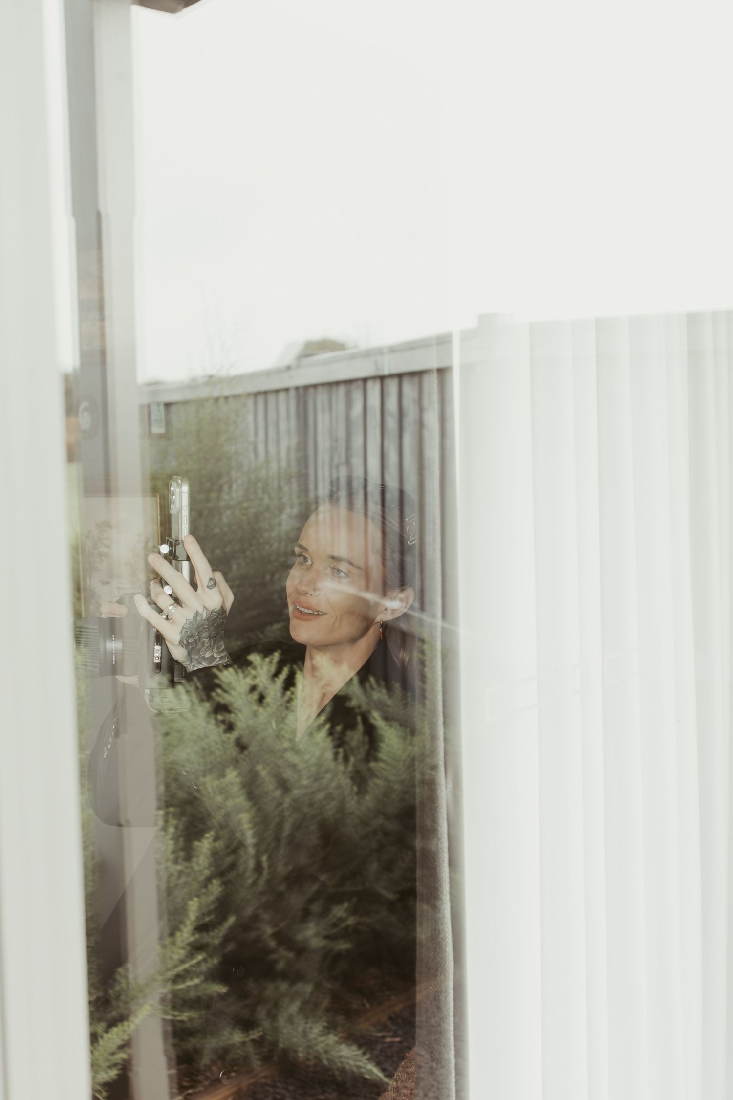 A smiling woman with tattooed hands takes a selfie through a glass window surrounded by greenery.