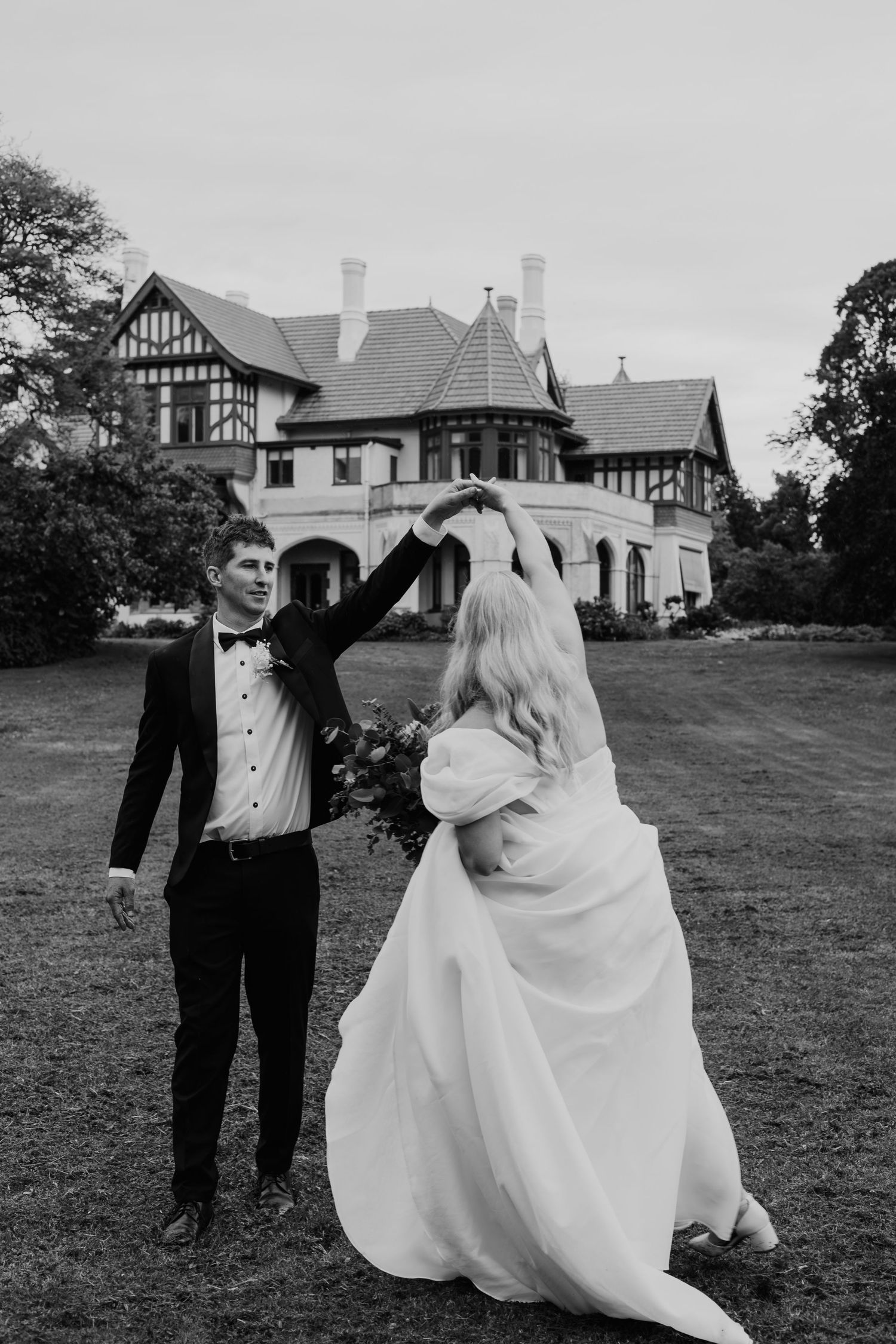 Black and white photo of groom spinning bride in flowing wedding gown on lawn before manor.