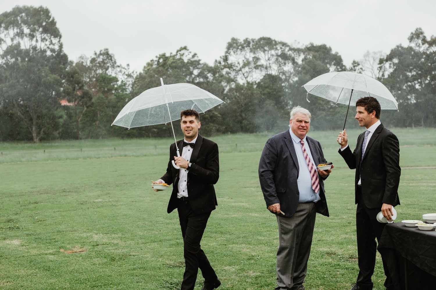 Three men in suits carrying clear umbrellas walk across misty green lawn at outdoor wedding.