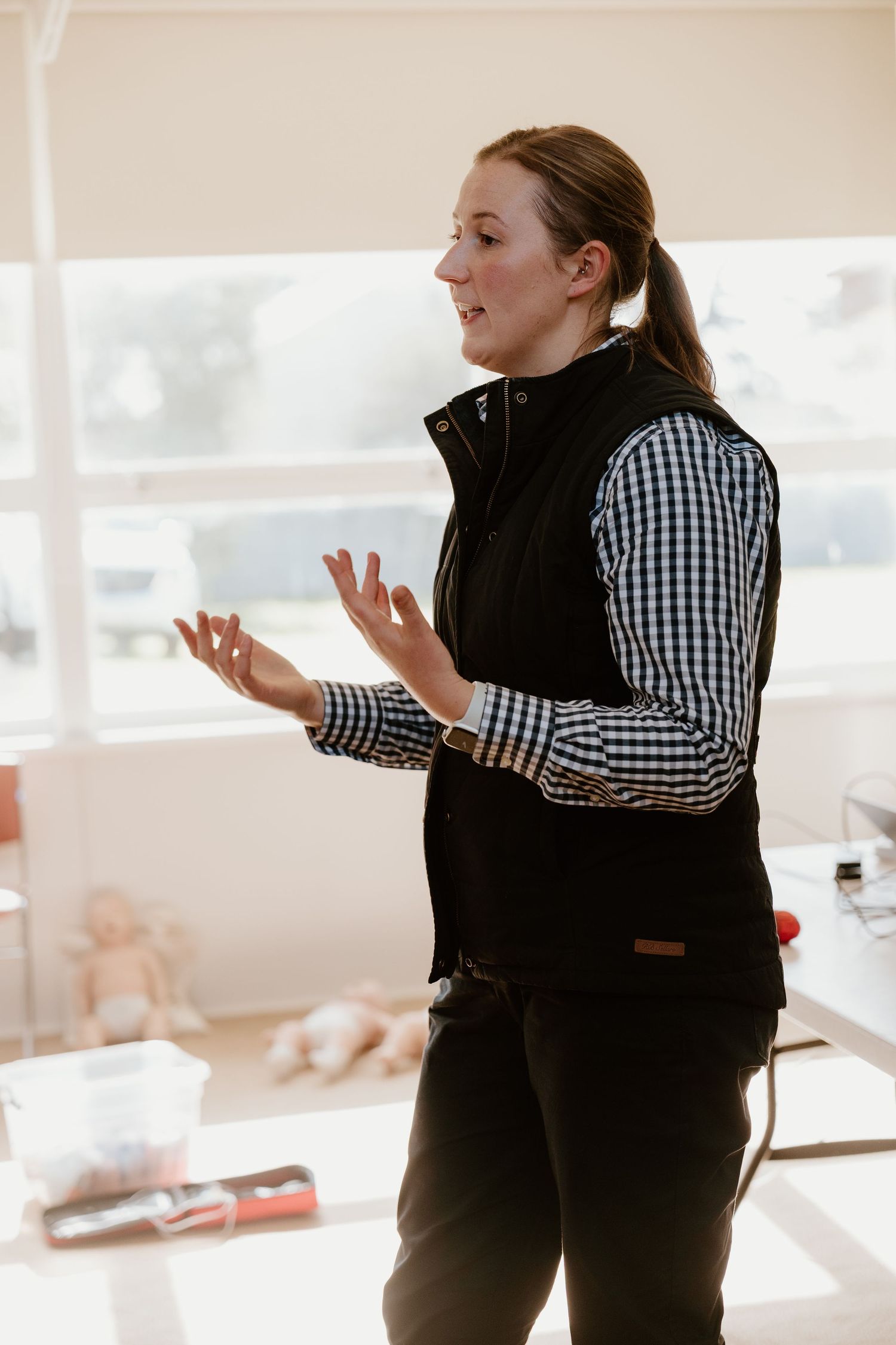 Woman in black vest and plaid shirt gesturing while speaking in a bright classroom setting.