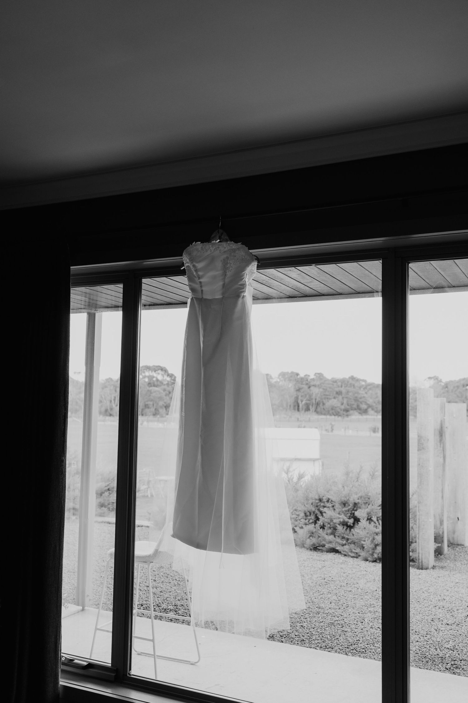 A wedding dress hanging in a window with a garden view, captured in black and white.