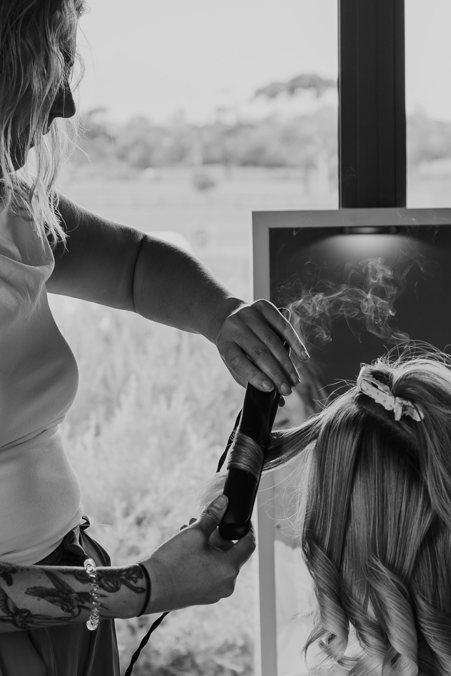 Black and white photo of a hairstylist curling a bride's hair with steam rising from the curling iron.