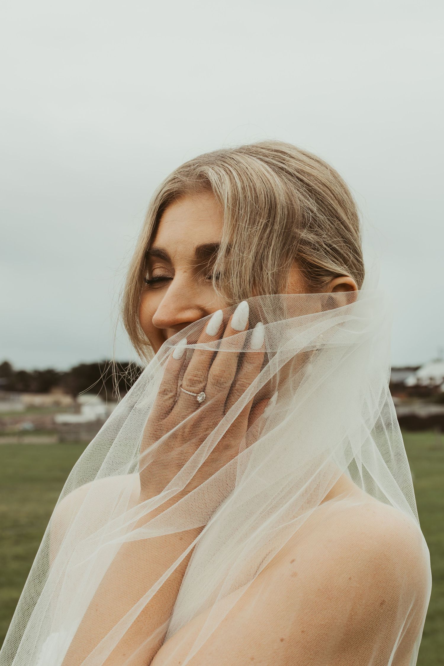 Bride with eyes closed holding sheer veil over face, displaying diamond ring against soft grey sky.