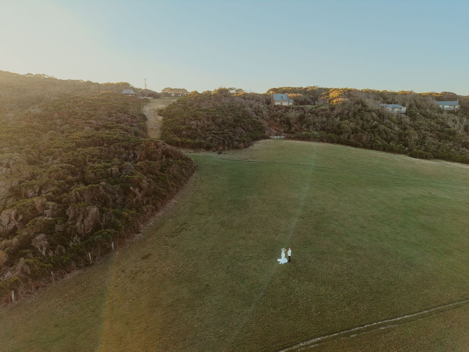 Aerial drone view of bride and groom on their wedding day in  a lush green field at golden hour with trees and coastal houses on a hillside.
