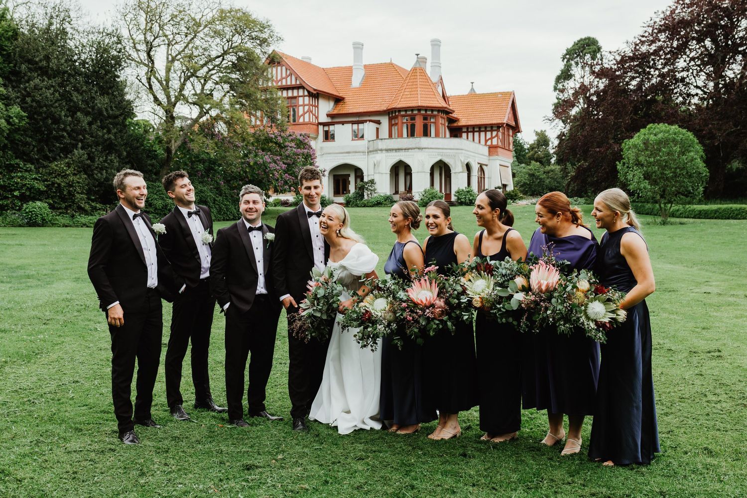 Wedding party in formal attire poses on green lawn before elegant manor house, bridesmaids holding large native floral bouquets.