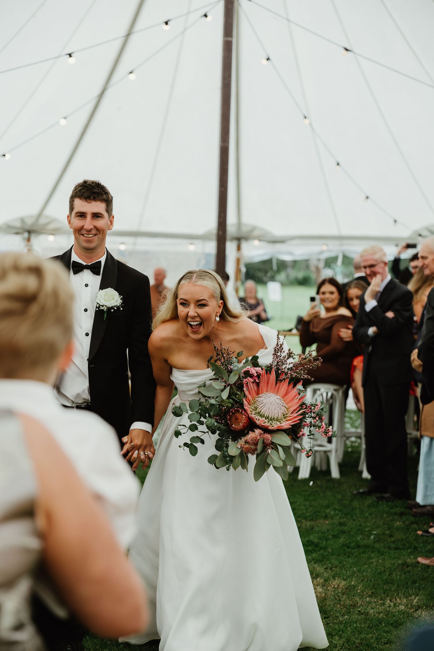 Joyful bride holding a king protea bouquet walks with groom down the aisle under a white tent at an outdoor wedding.
