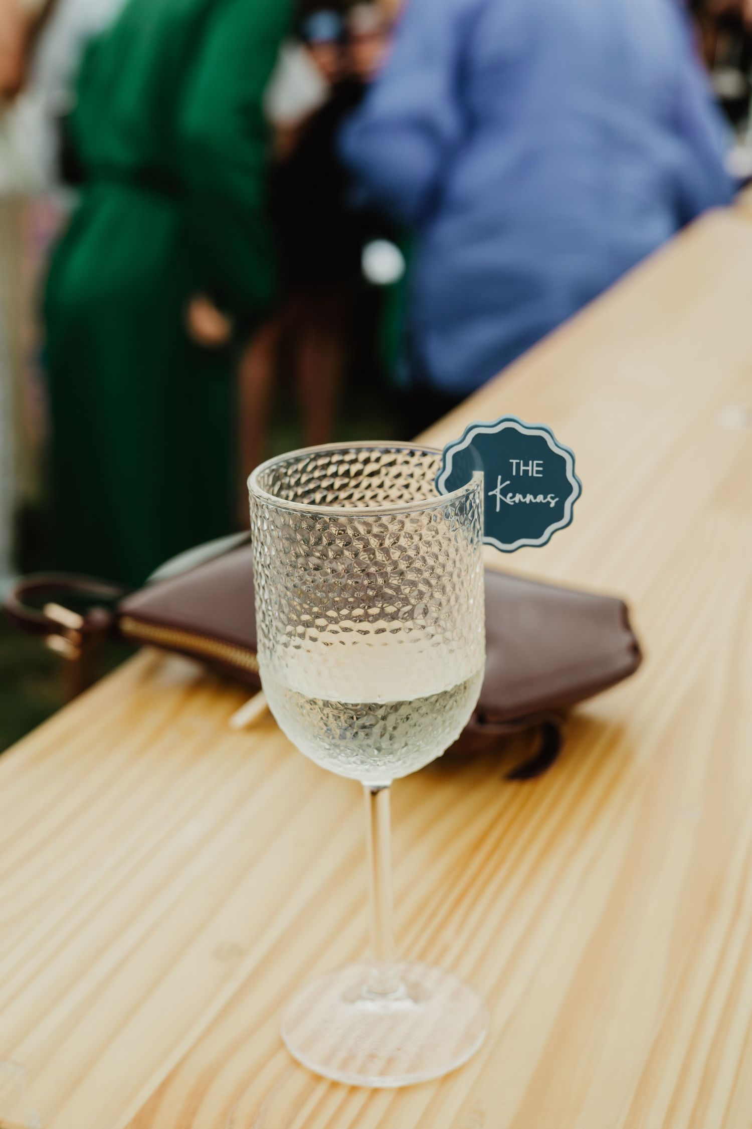 Glass of white wine with decorative tag reading The Kennas on wooden bar at wedding reception.
