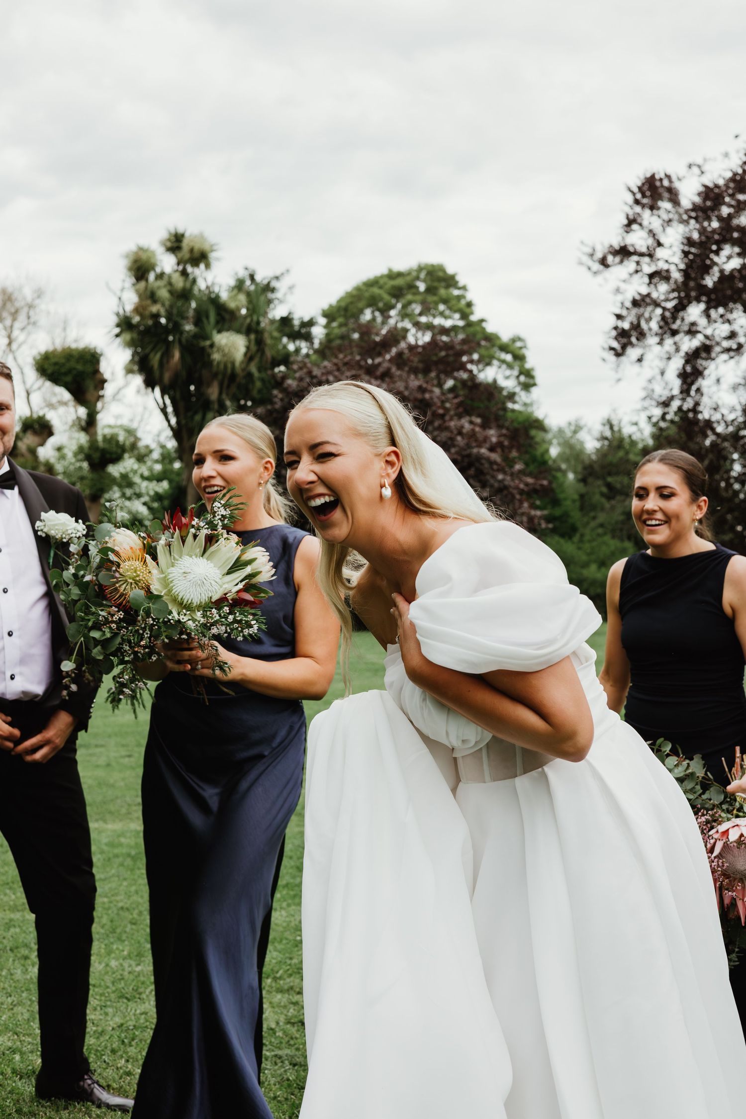 Laughing bride in off-shoulder white gown with bridesmaids holding bouquet outdoors.