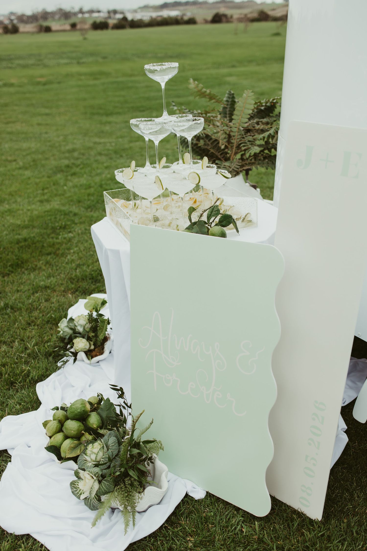 Elegant outdoor wedding champagne tower display with sage green signage and white floral arrangements.