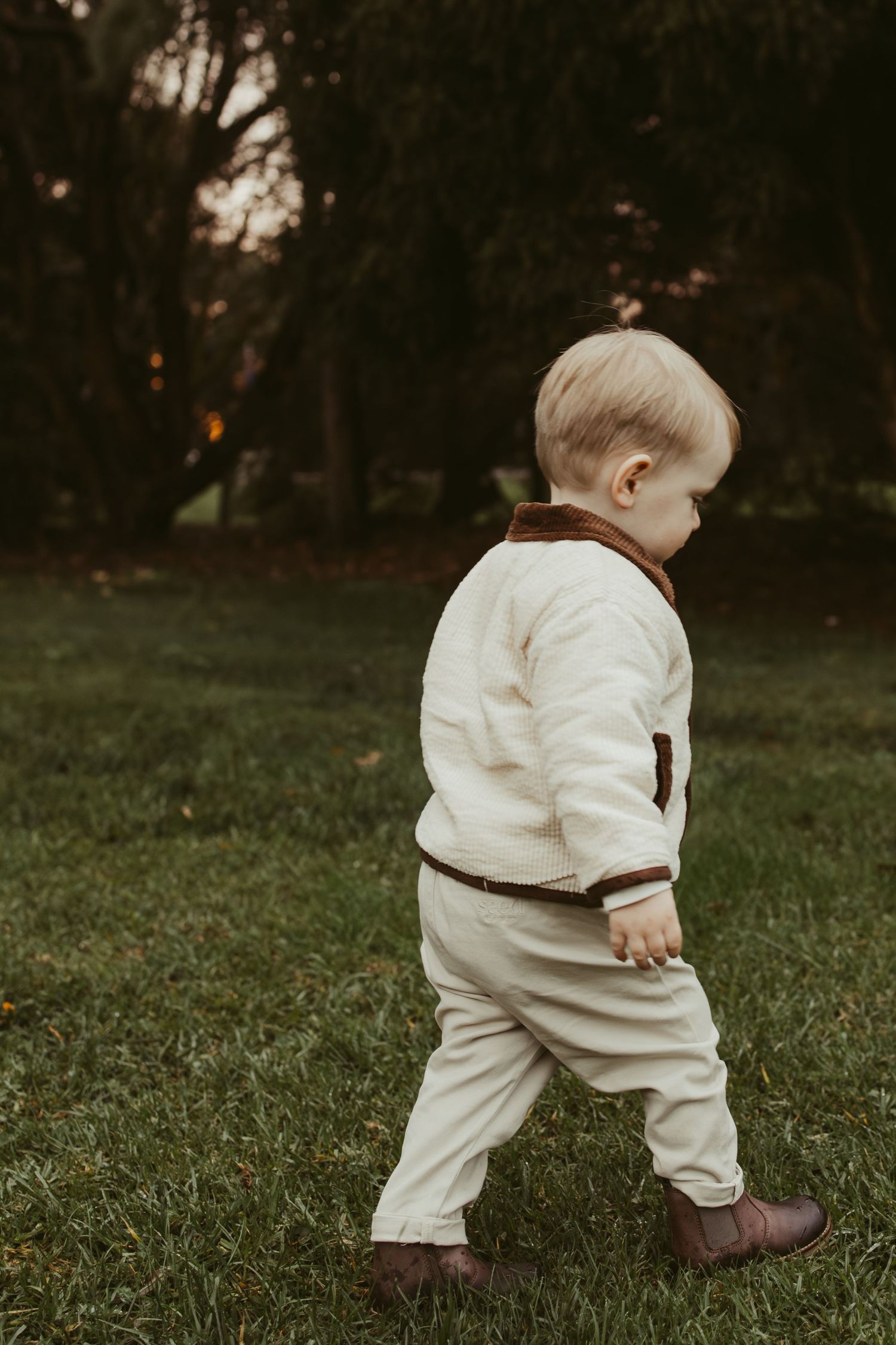 Adorable toddler in cream and brown outfit walking on green grass in a park.