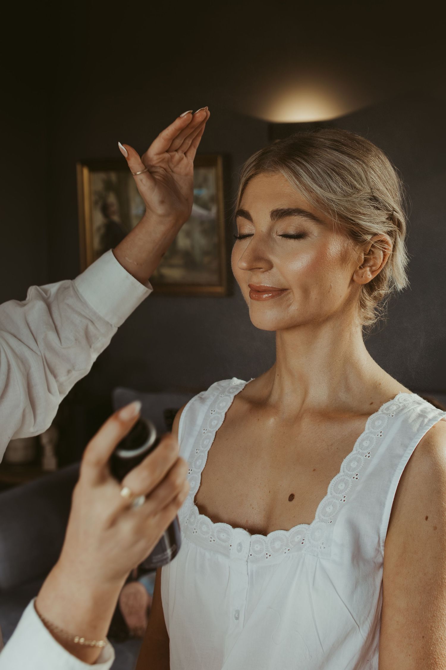 Bride getting makeup applied by artist, eyes closed, wearing white dress in elegant room.