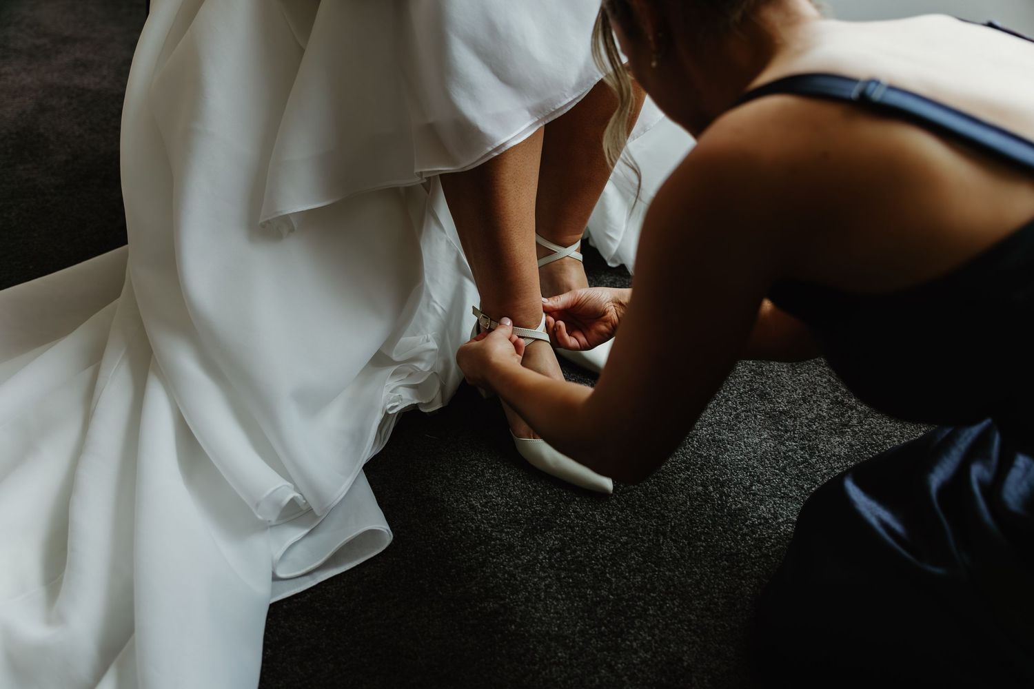 Bridesmaid helping bride put on white wedding shoes while kneeling on gray carpet in bridal suite.