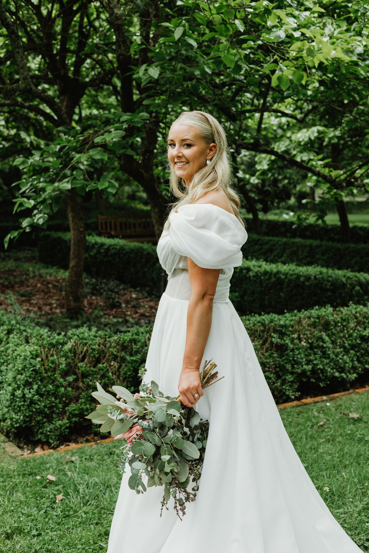 Smiling bride in off-shoulder white gown holds greenery bouquet in lush garden.