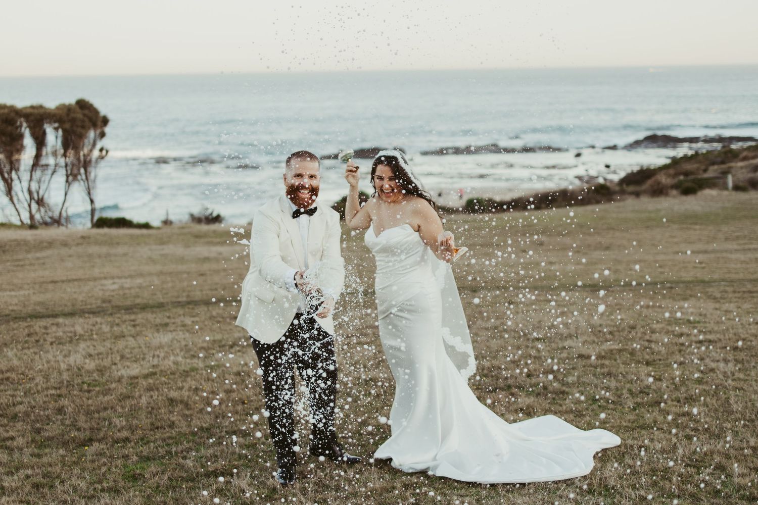 Bride and groom celebrating on coastal lawn with confetti, ocean backdrop, white wedding dress and suit.