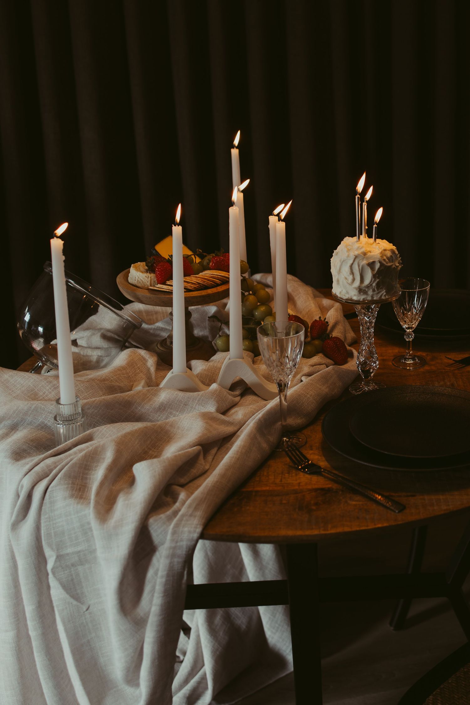 Elegant candlelit wedding table setting with white taper candles, linen runner, crystal glasses, and fruit display on rustic wood table.