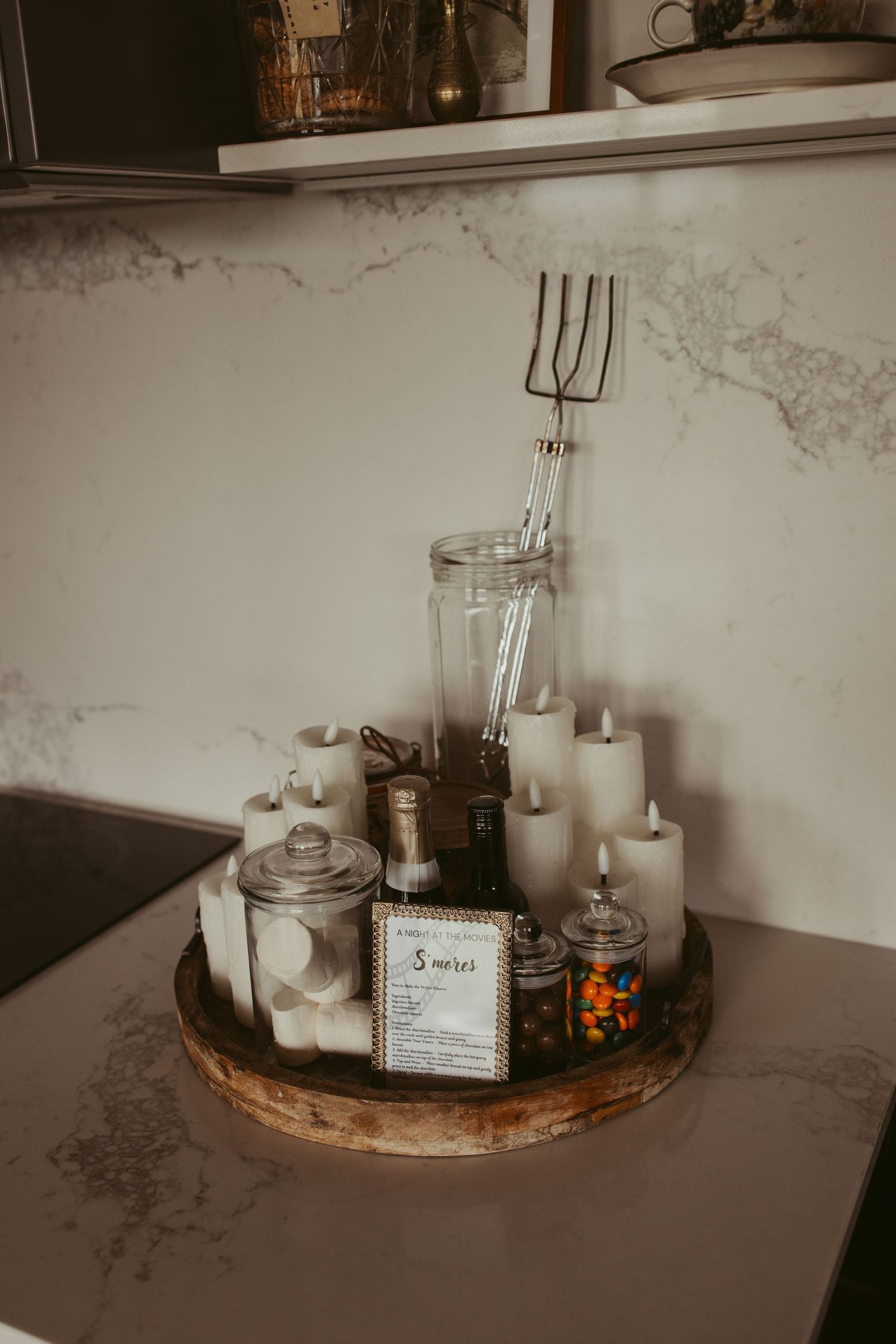 Rustic wooden tray with white candles, glass jars, and decorative items on marble kitchen counter.