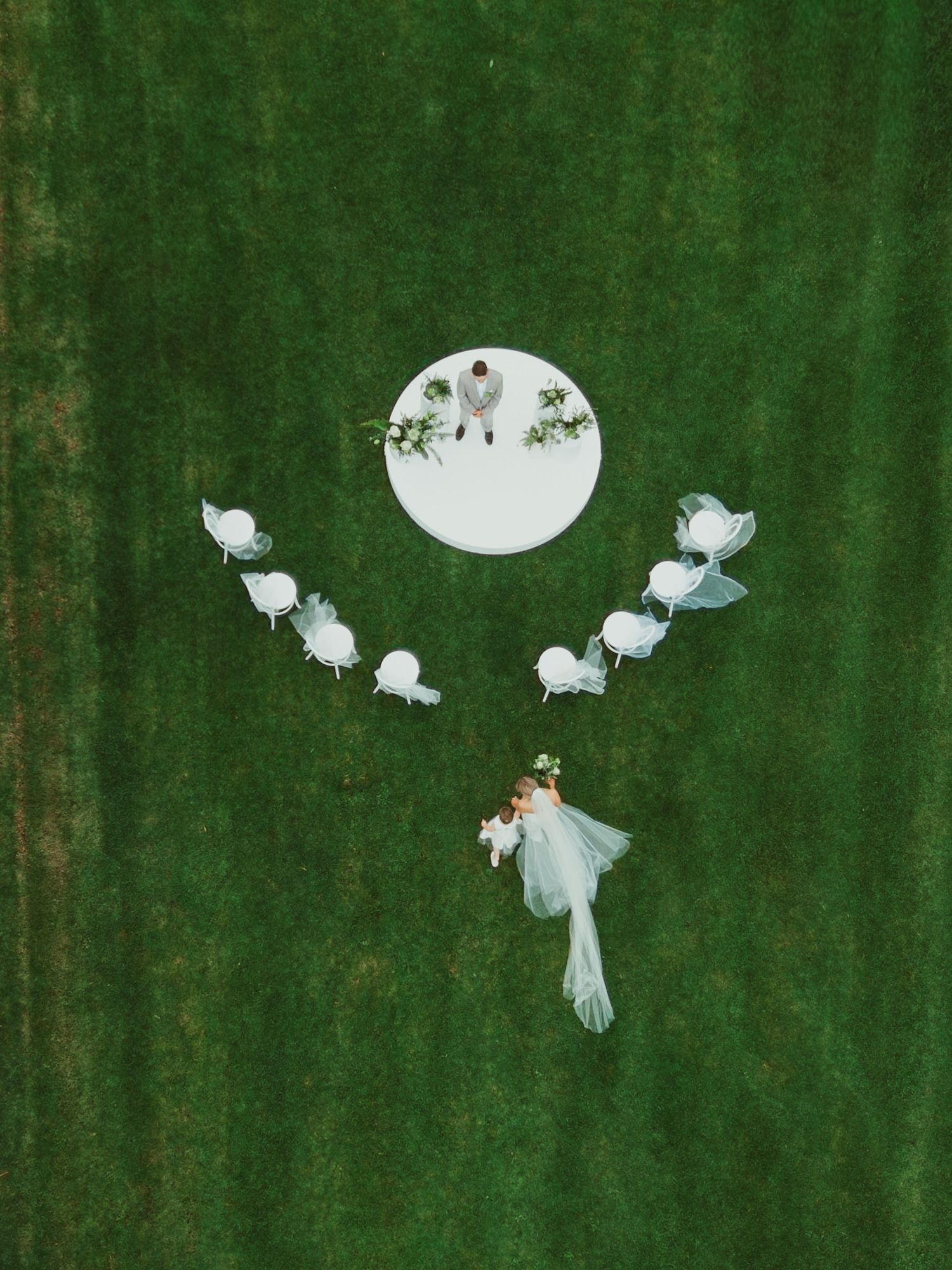 Aerial drone view of a wedding ceremony on a white circular platform surrounded by white chairs on a lush green lawn.