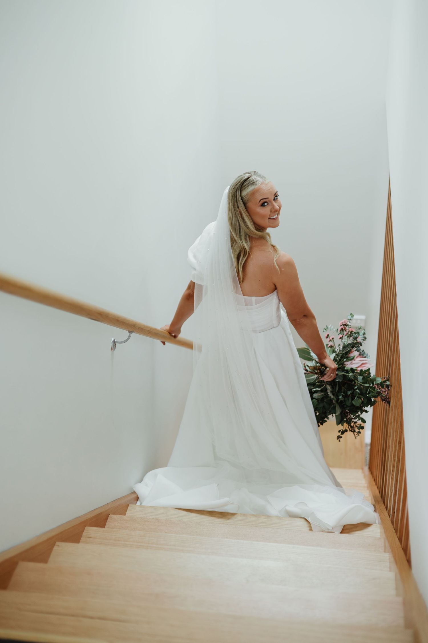 Elegant bride in white gown holding bouquet pauses on wooden staircase wearing cathedral veil.