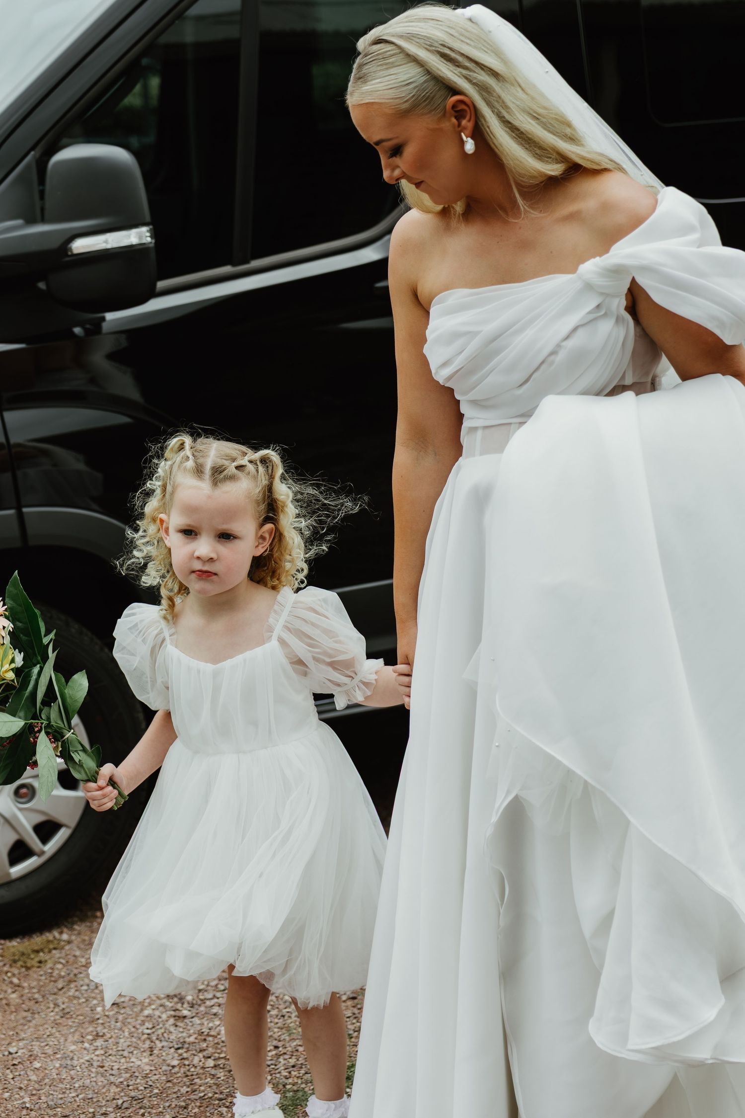 Bride in white off-shoulder gown holds hands with flower girl in white dress beside black vehicle.