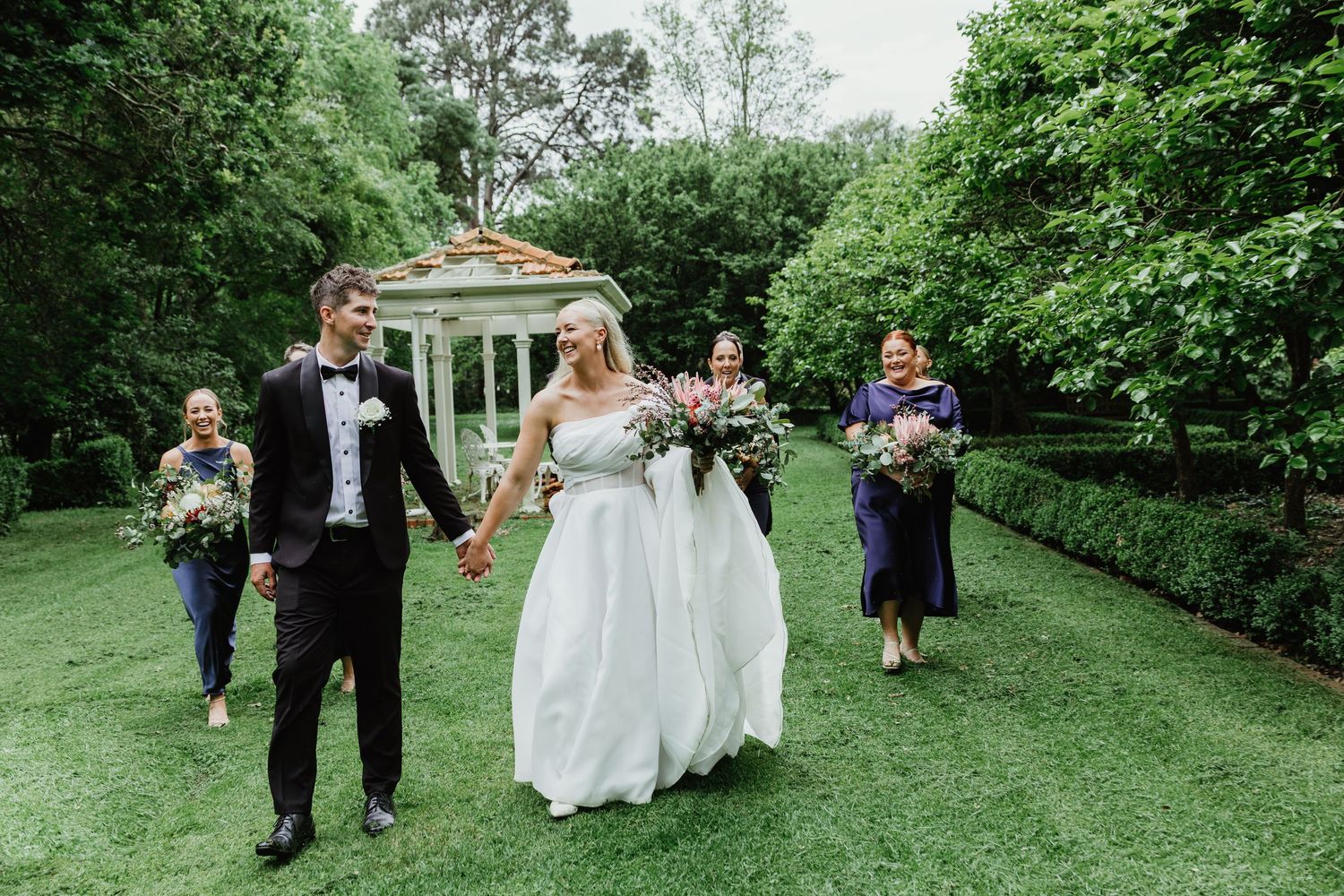Bride and groom walking hand in hand with bridesmaids in navy dresses in garden.