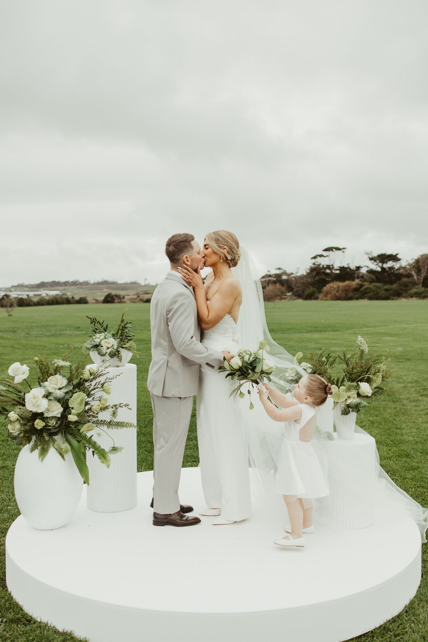Bride and groom kissing on white circular altar with flower girl and green floral arrangements.