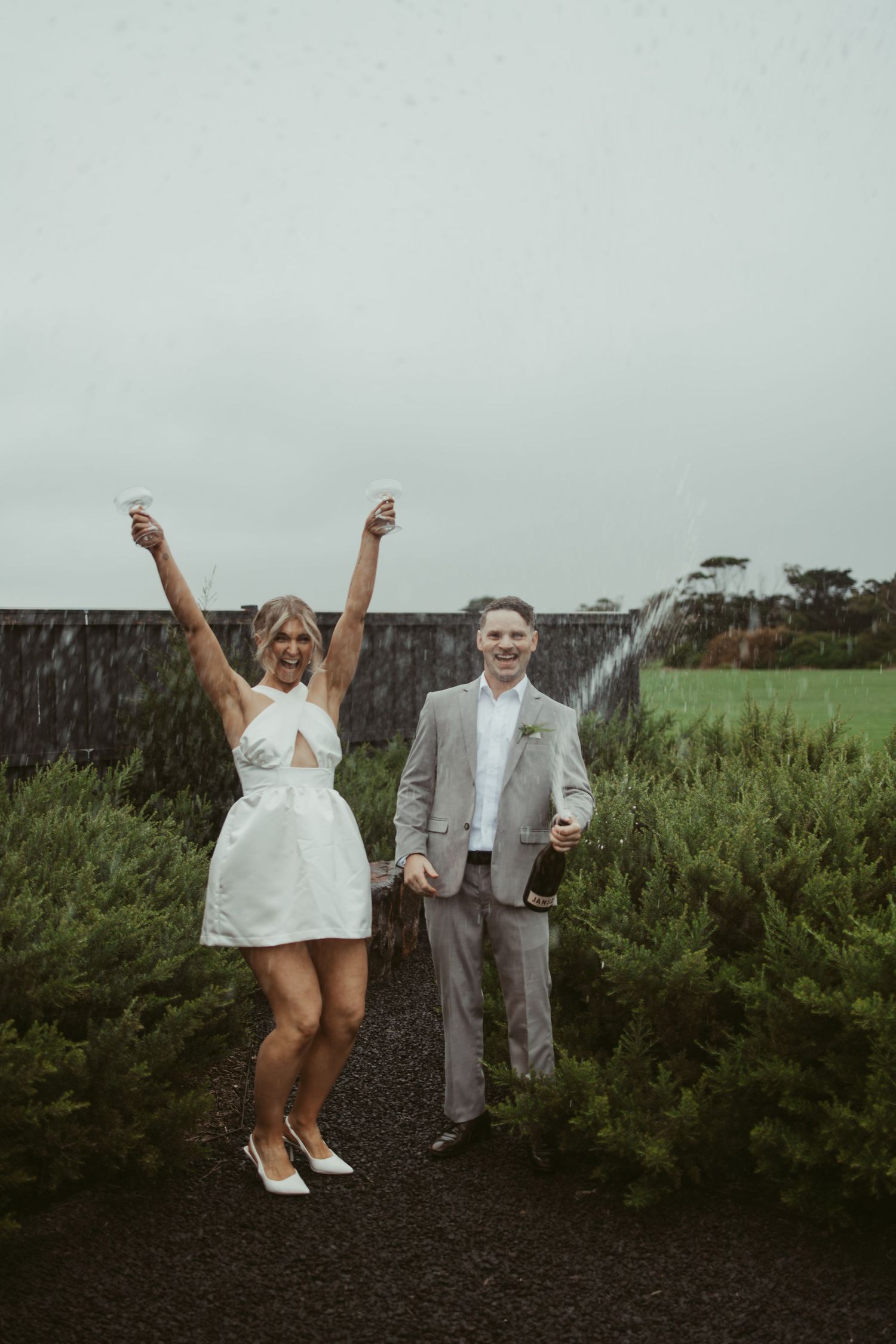 Happy bride and groom celebrating in the rain, bride raising arms with drinks on their wedding day.