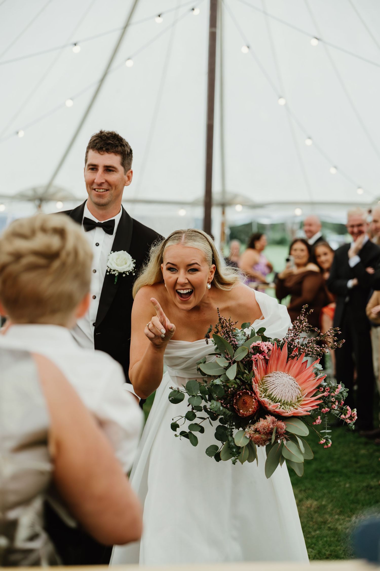 Joyful bride pointing and laughing with groom during wedding reception under a white sailcloth tent.