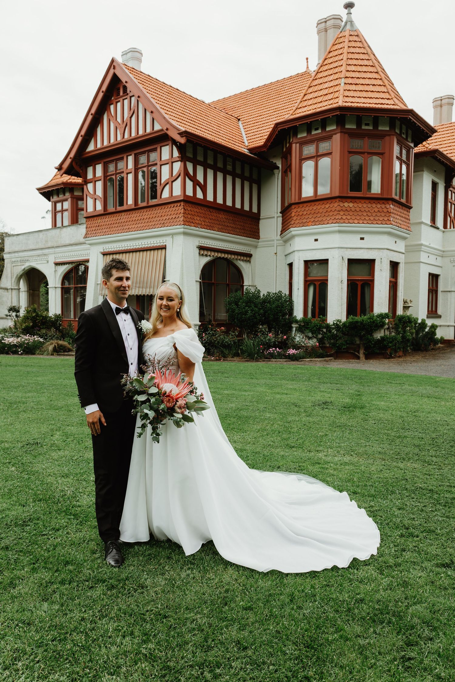 Bride and groom pose on green lawn before grand Tudor-style mansion on their wedding day.