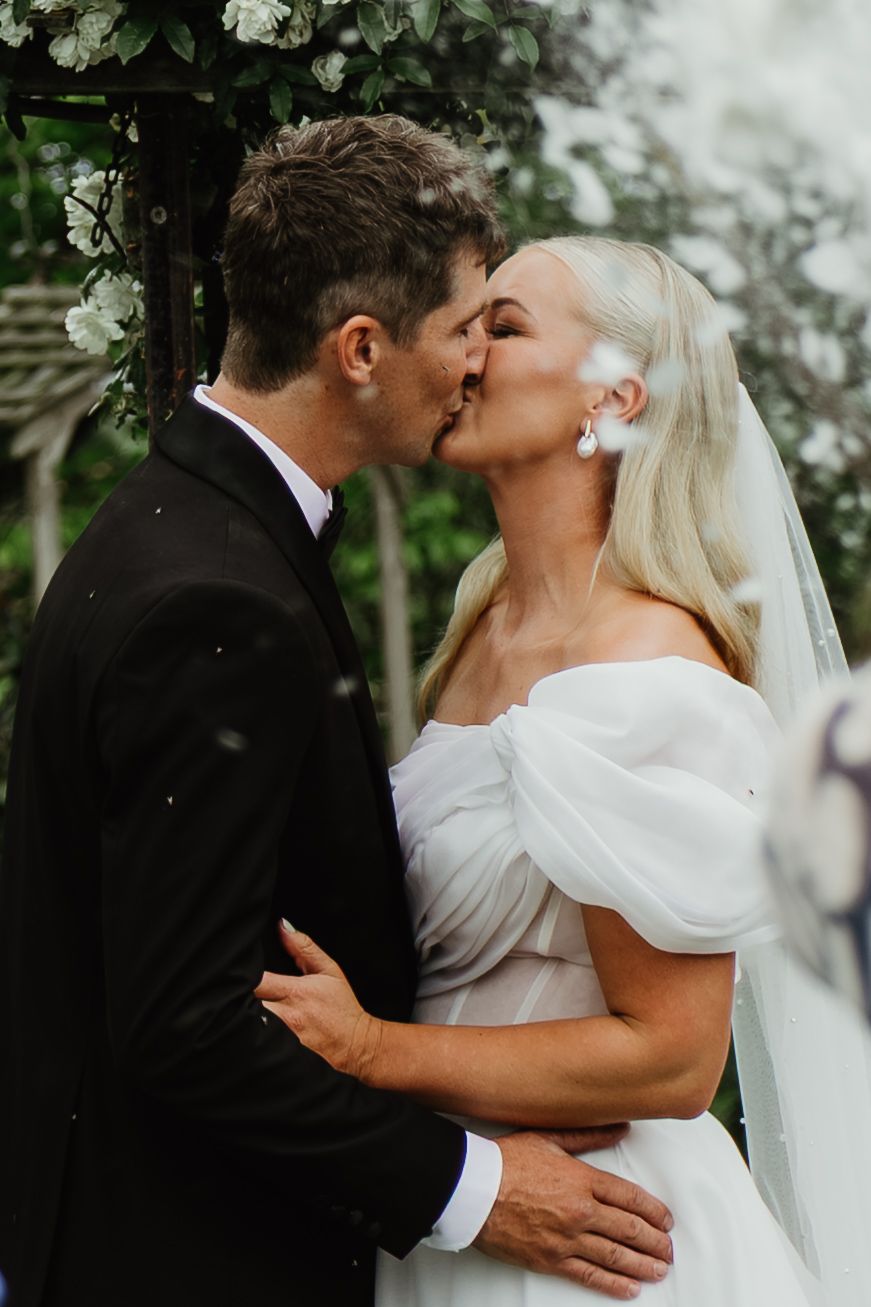 Bride and groom share their first kiss under a floral arch, bride wearing an off-shoulder white gown.