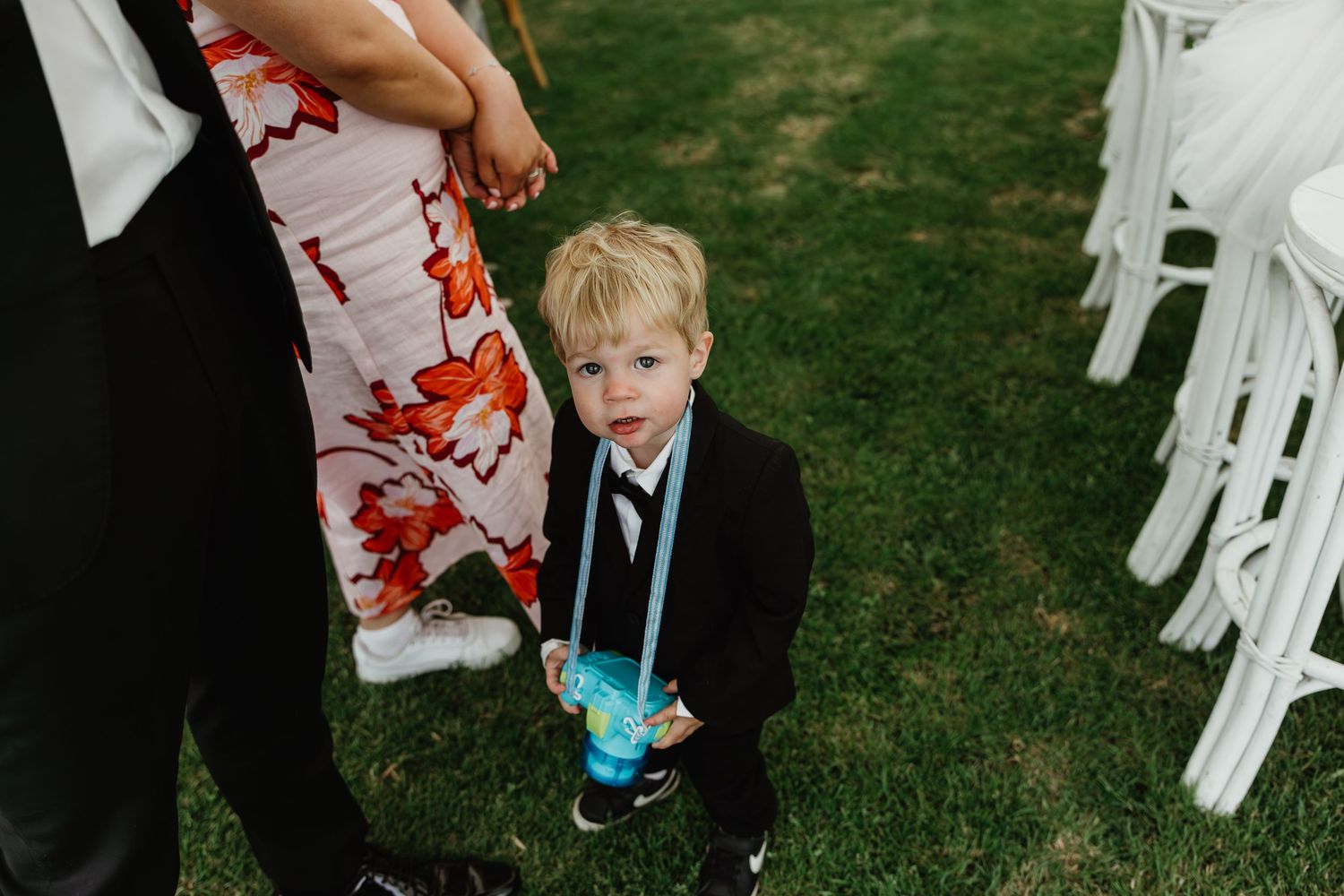 Young blond boy in black suit holding drink at outdoor wedding ceremony.