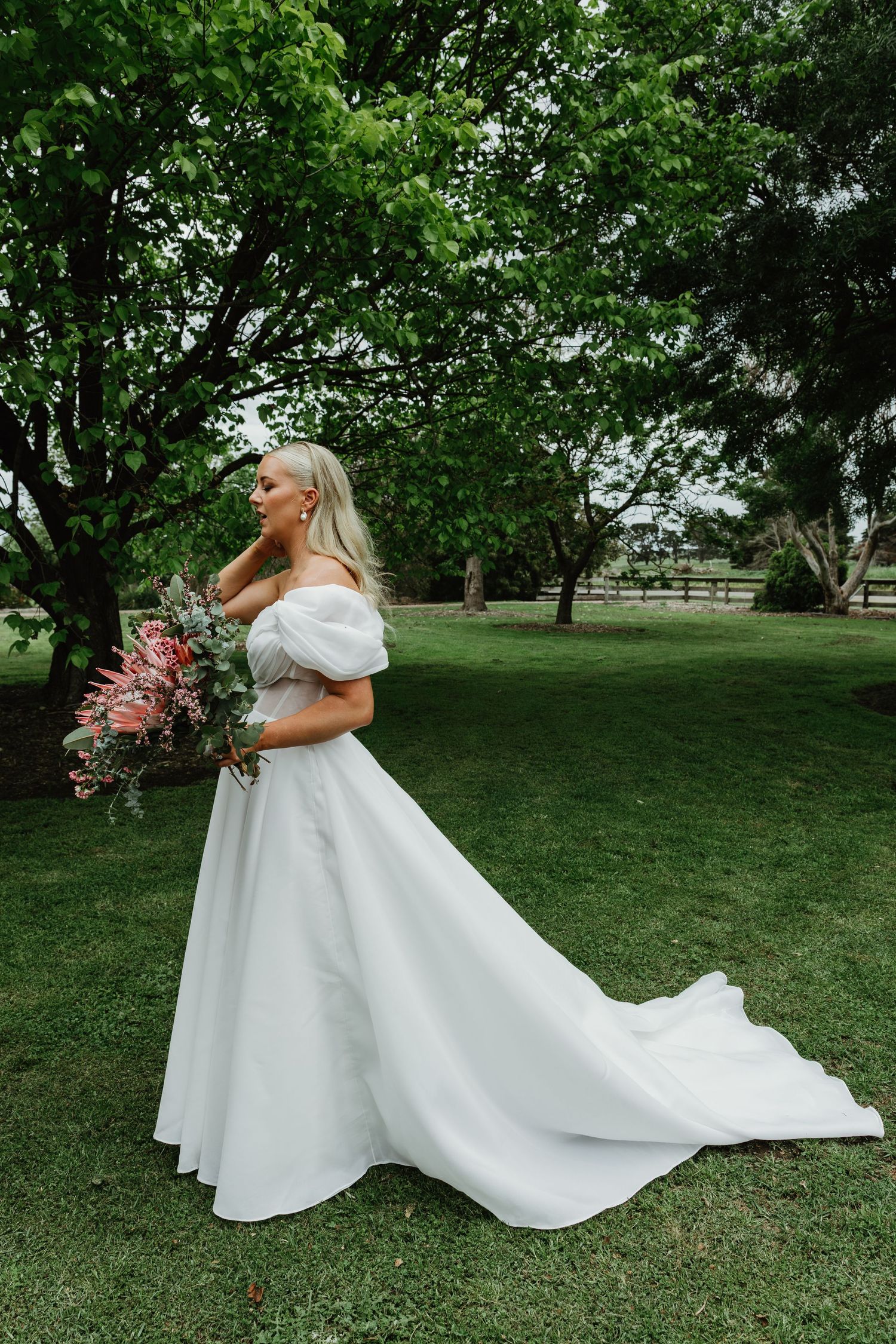 Bride in off-shoulder white ball gown holding pink bouquet standing under green trees.