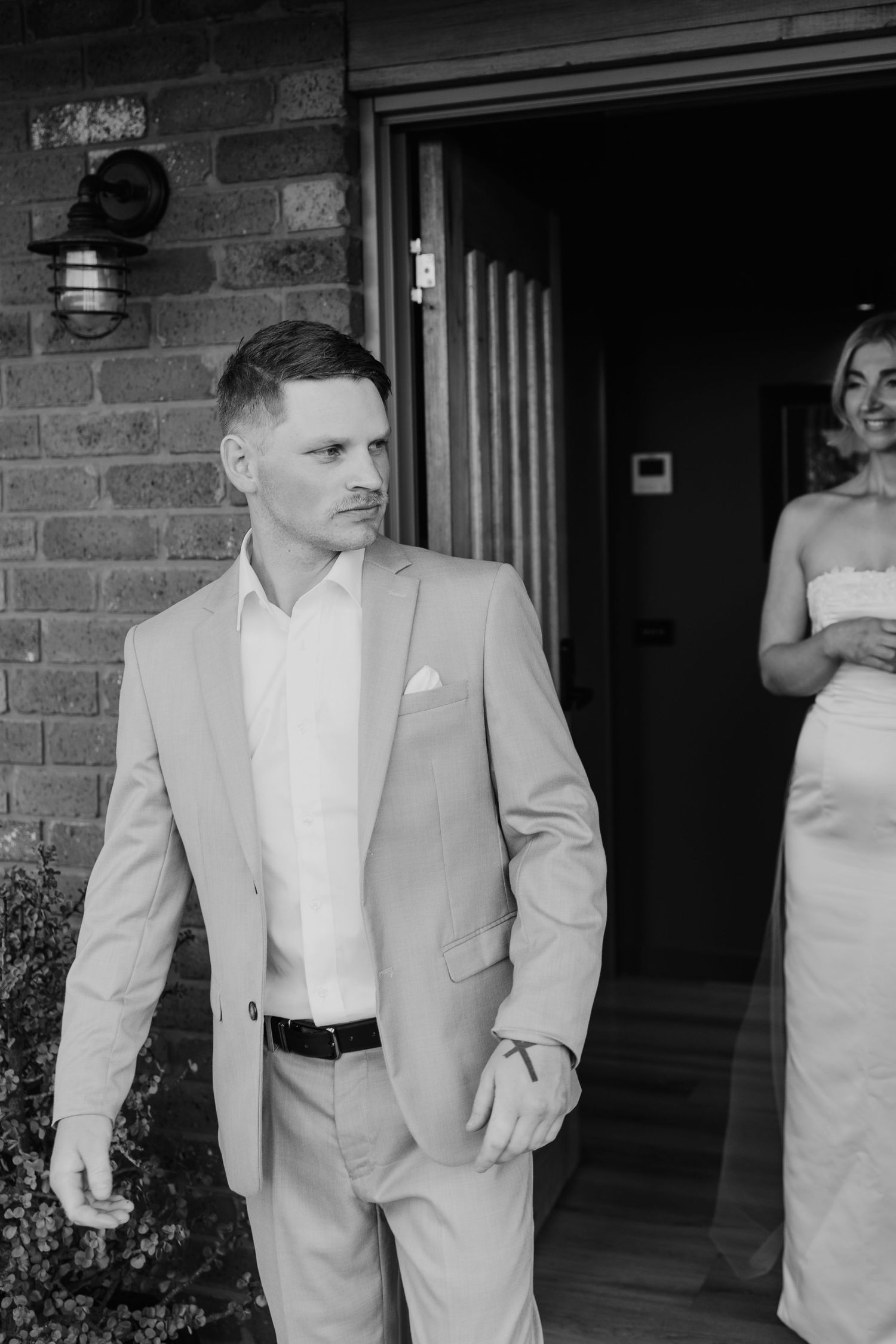 Black and white photo of groom in grey suit stepping outside with bride behind.
