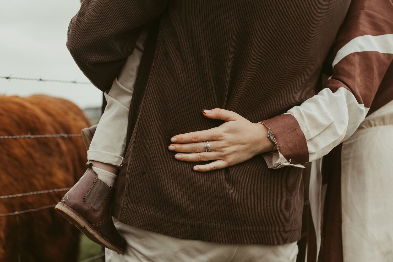 Woman in brown and white dress embracing man near a Highland cow farm fence.