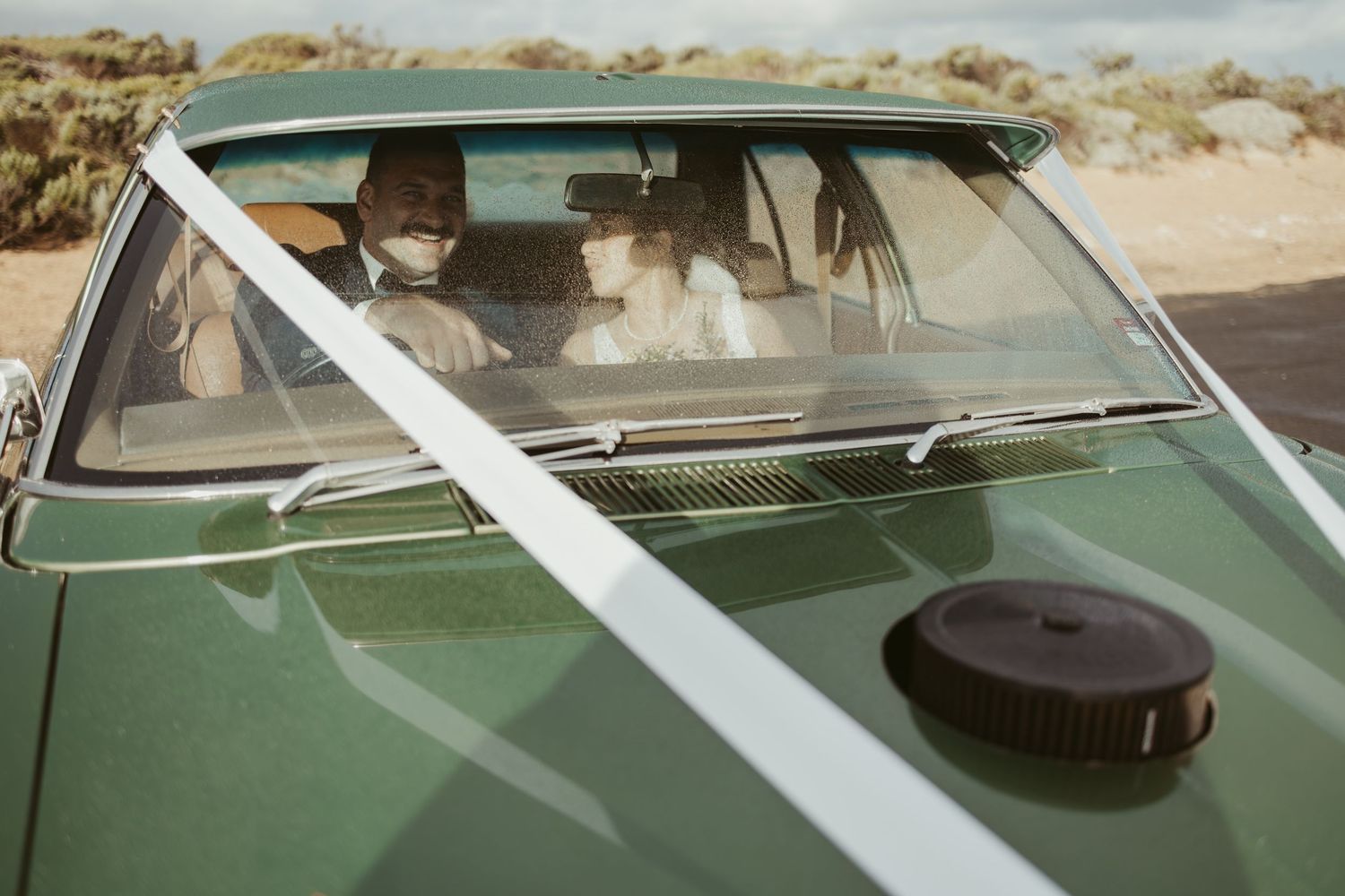 Couple in a vintage green convertible car decorated with white ribbon, parked in a desert landscape.