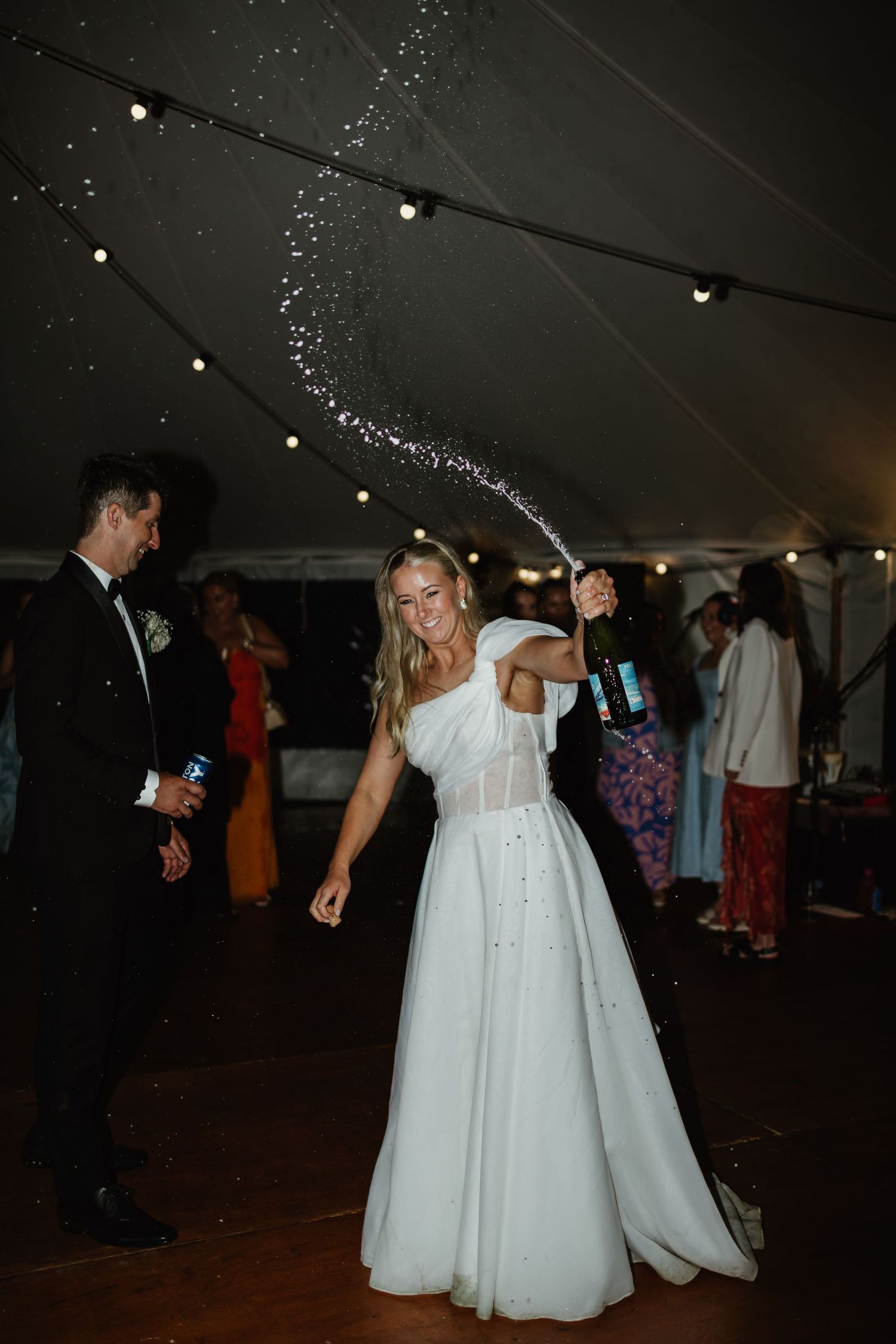 Joyful bride spraying champagne on the dance floor during lively wedding reception under a marquee tent.
