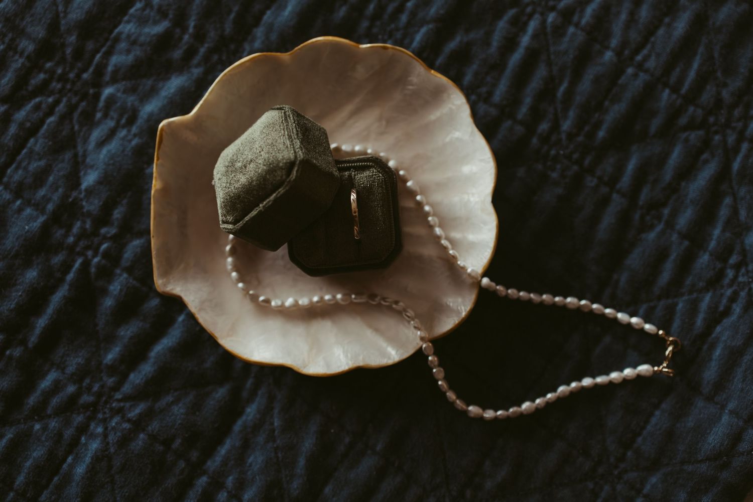 A pearl necklace and dark velvet ring boxes rest in a decorative shell dish on a quilted navy fabric.