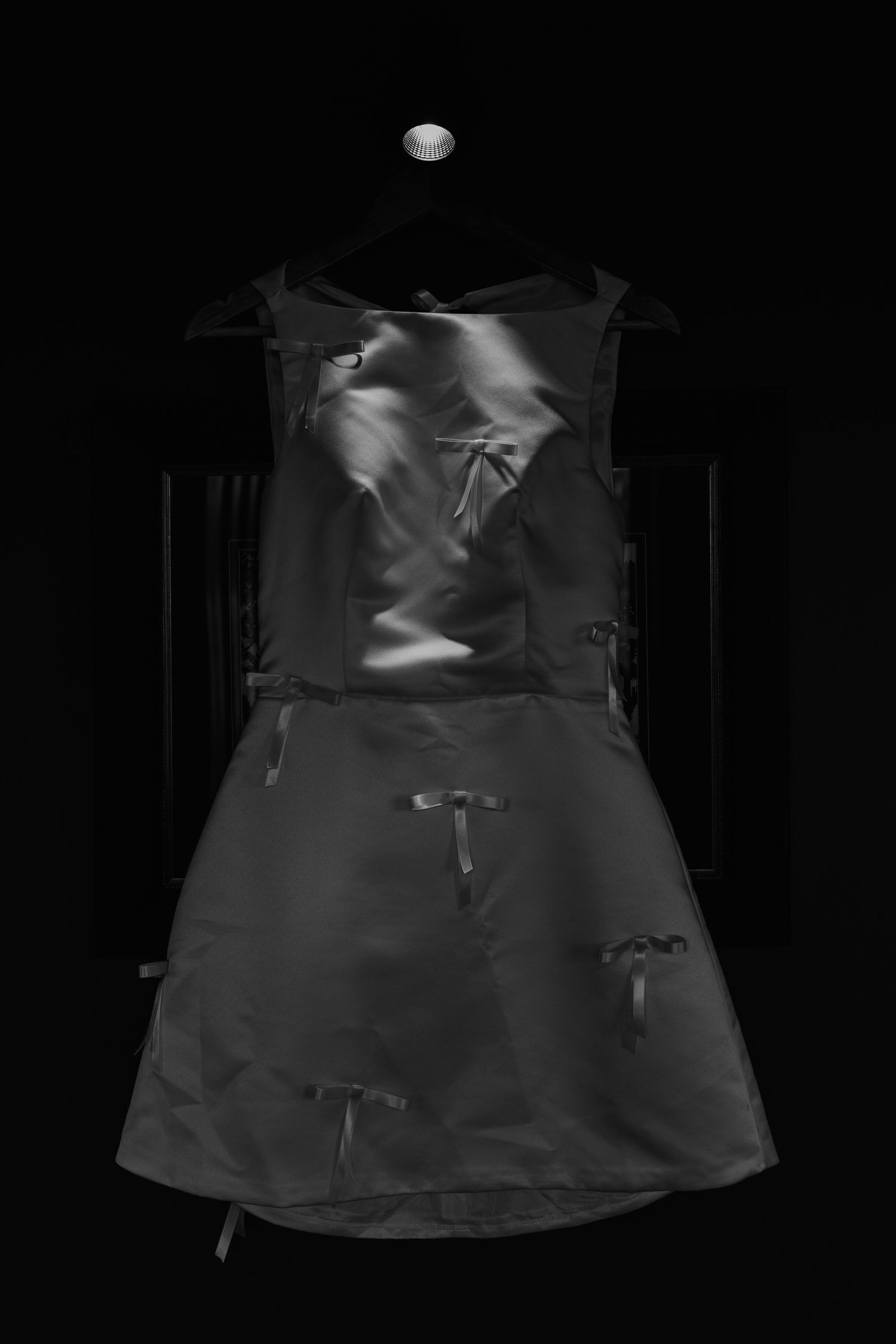 Black and white photo of an elegant sleeveless dress with bow details hanging on a hanger against dark background.