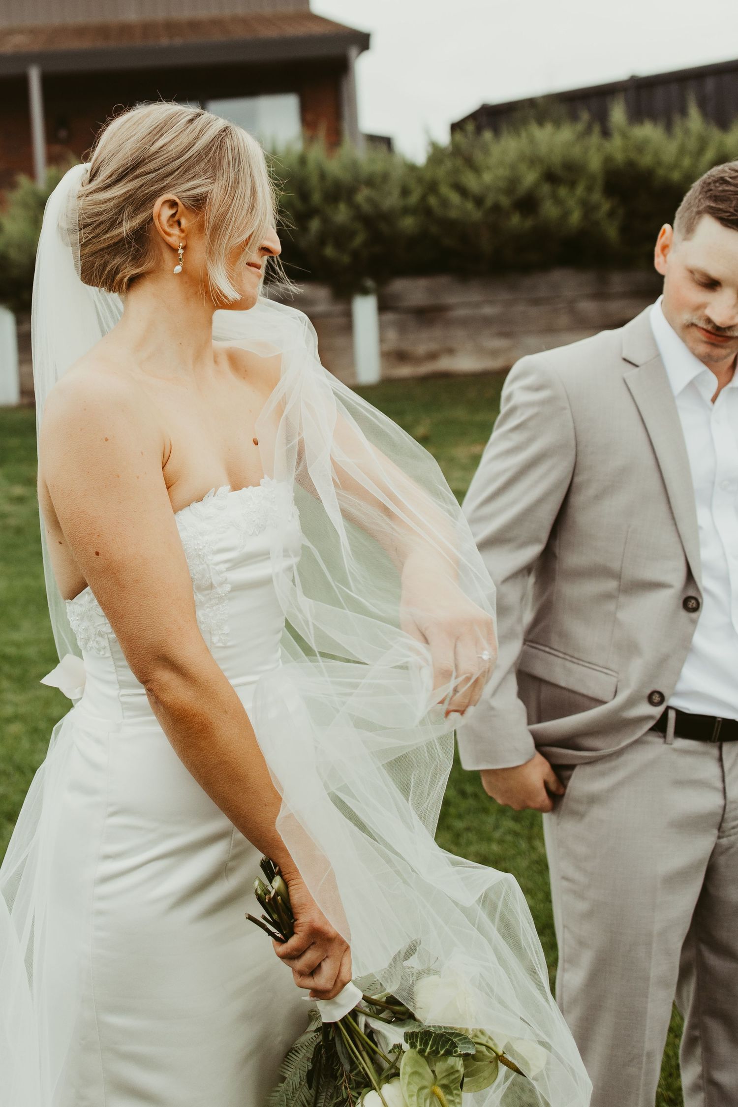 Bride in strapless white gown with flowing veil holds bouquet while walking alongside groom in grey suit.