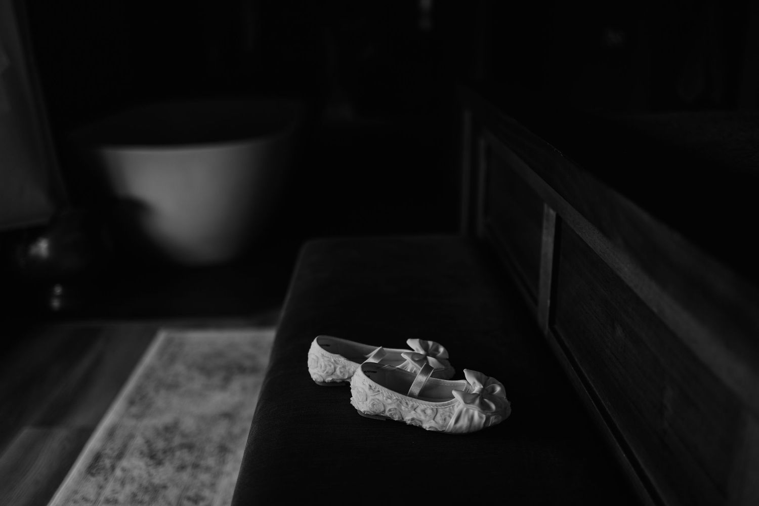 Black and white photo of delicate white bridal shoes with bow detail resting on a dark bench.