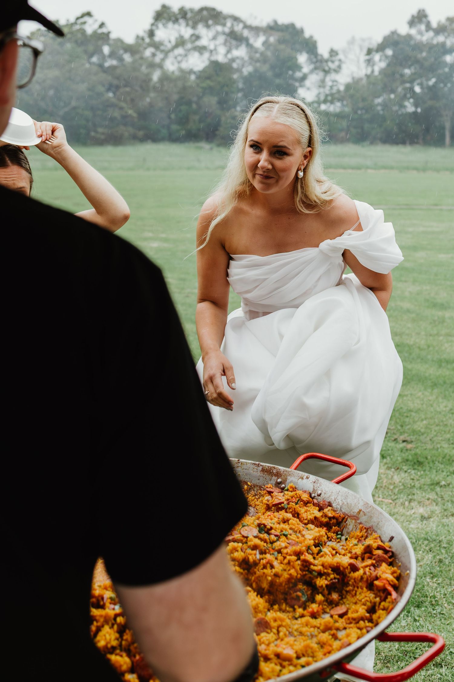 Bride in off-shoulder white gown smiles beside large paella pan at outdoor wedding reception.