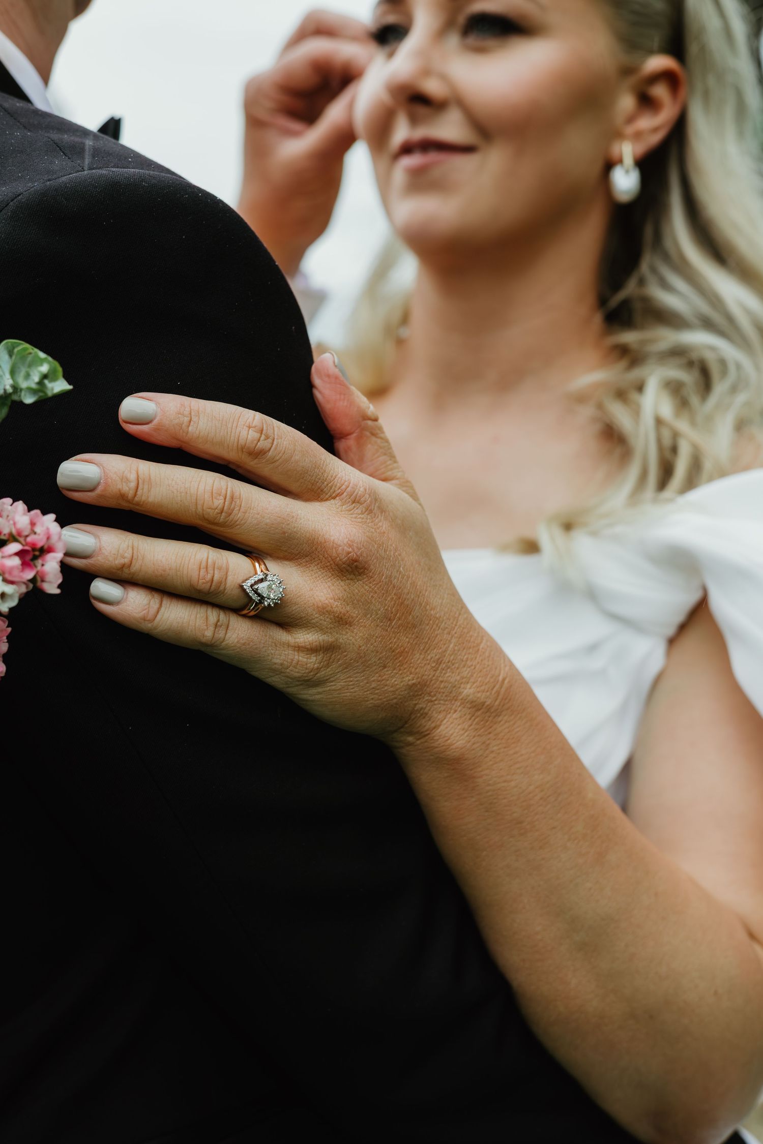 Close-up of bride's diamond wedding ring as she embraces groom in black tuxedo.