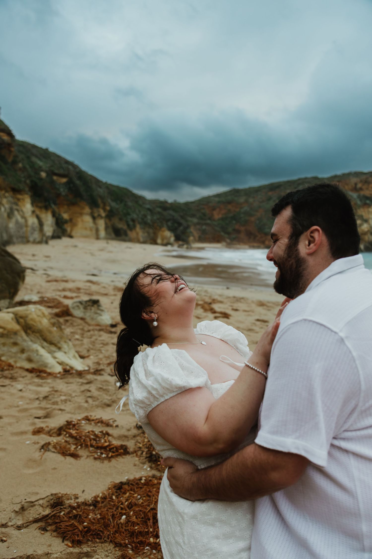 Couple embracing and laughing on a beach with dramatic rocky cliffs and stormy sky behind them.
